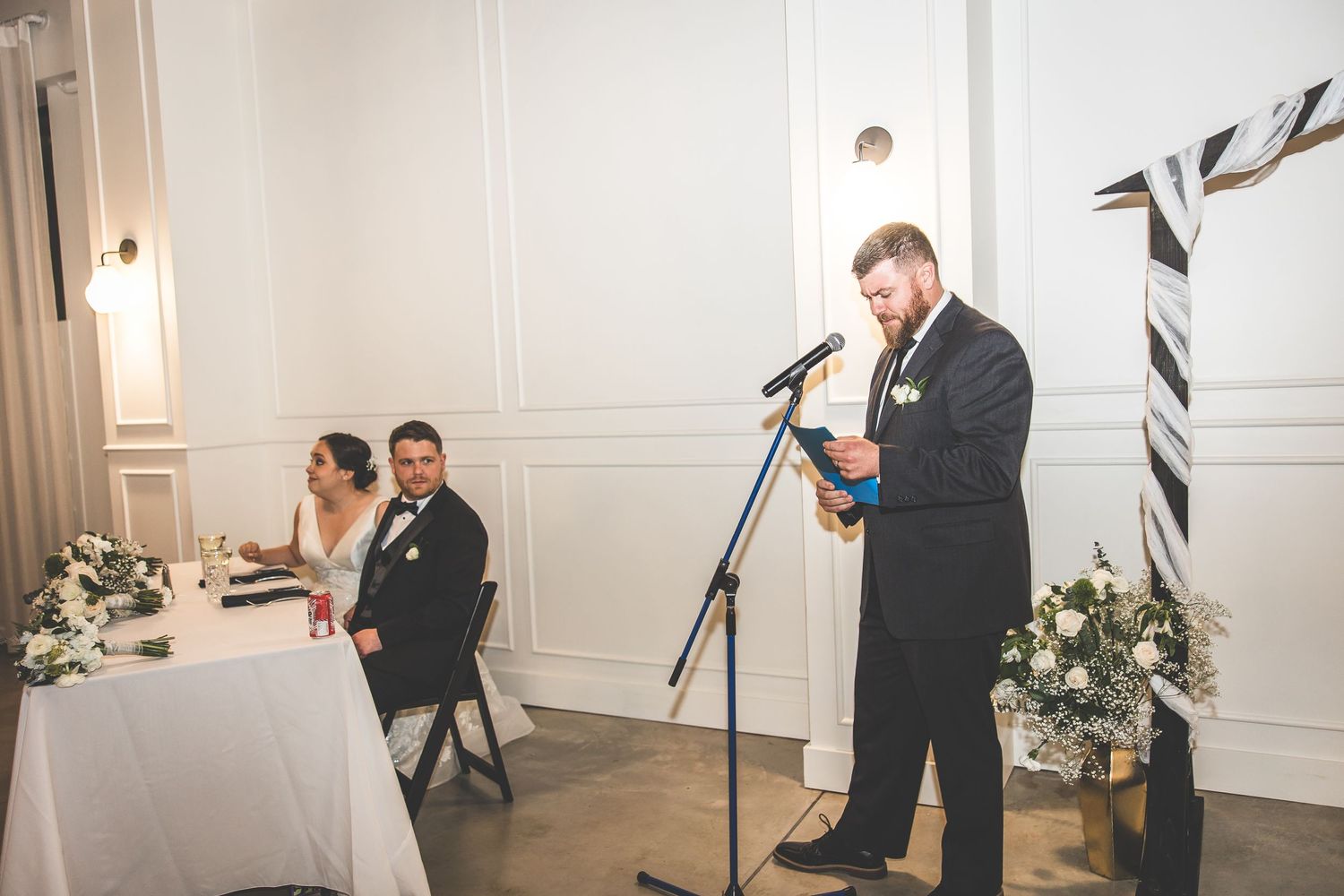 A speech being delivered at a wedding reception with decorative floral arrangements on the head table.