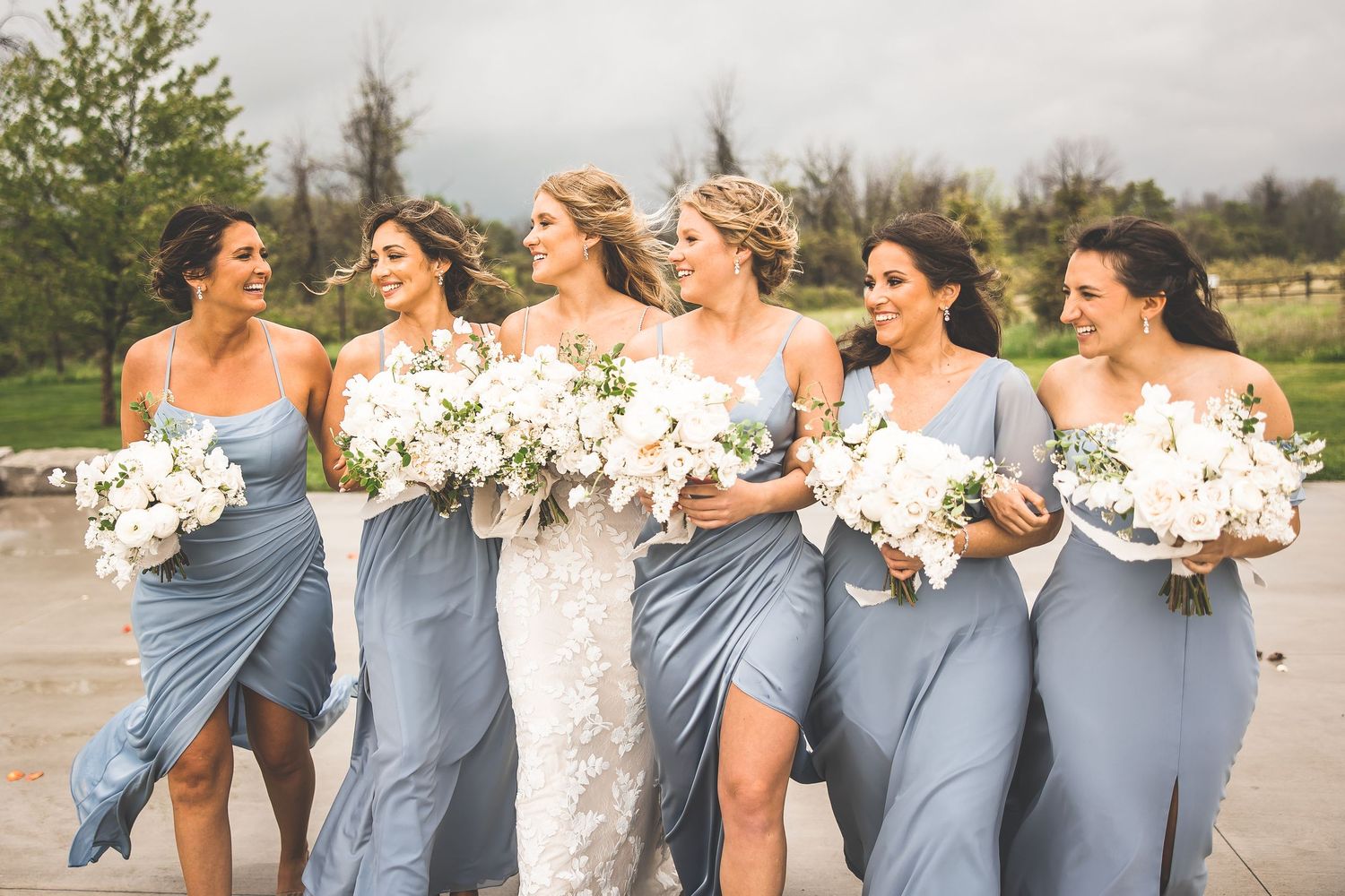 Bridal party in flowing blue dresses poses with white bouquets against cloudy sky.
