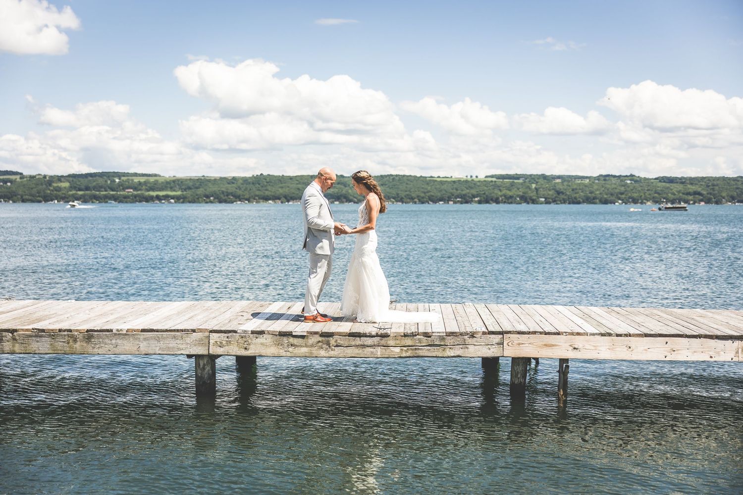 A wedding couple stands together on a wooden dock overlooking a serene lake.