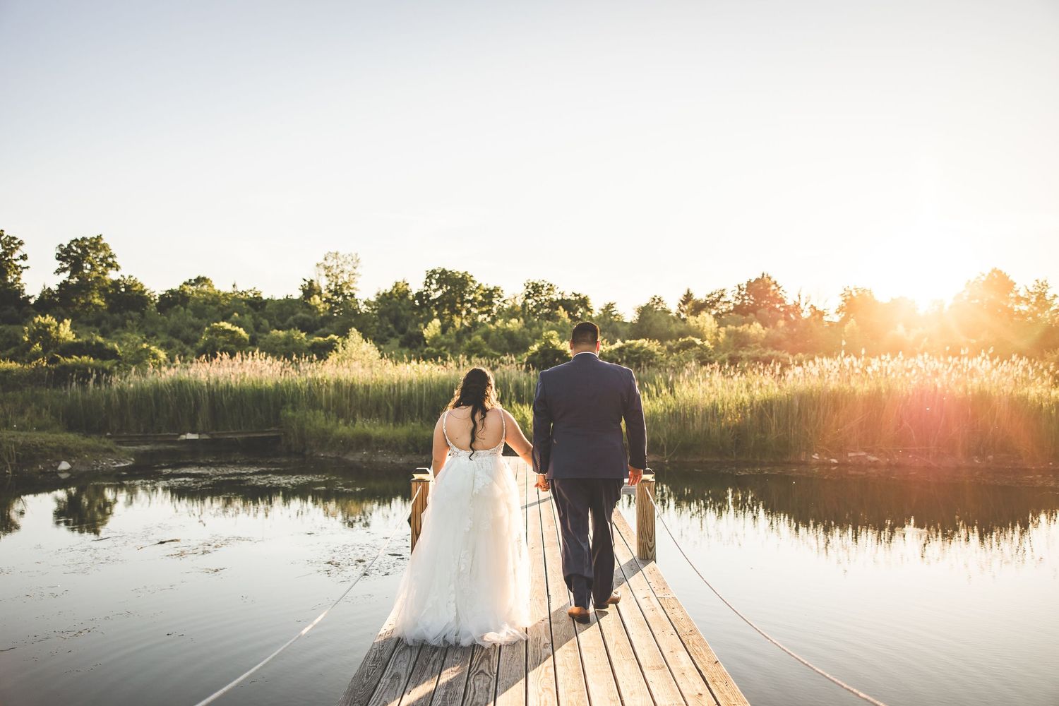 Wedding couple poses on wooden boardwalk against sunset lake backdrop.