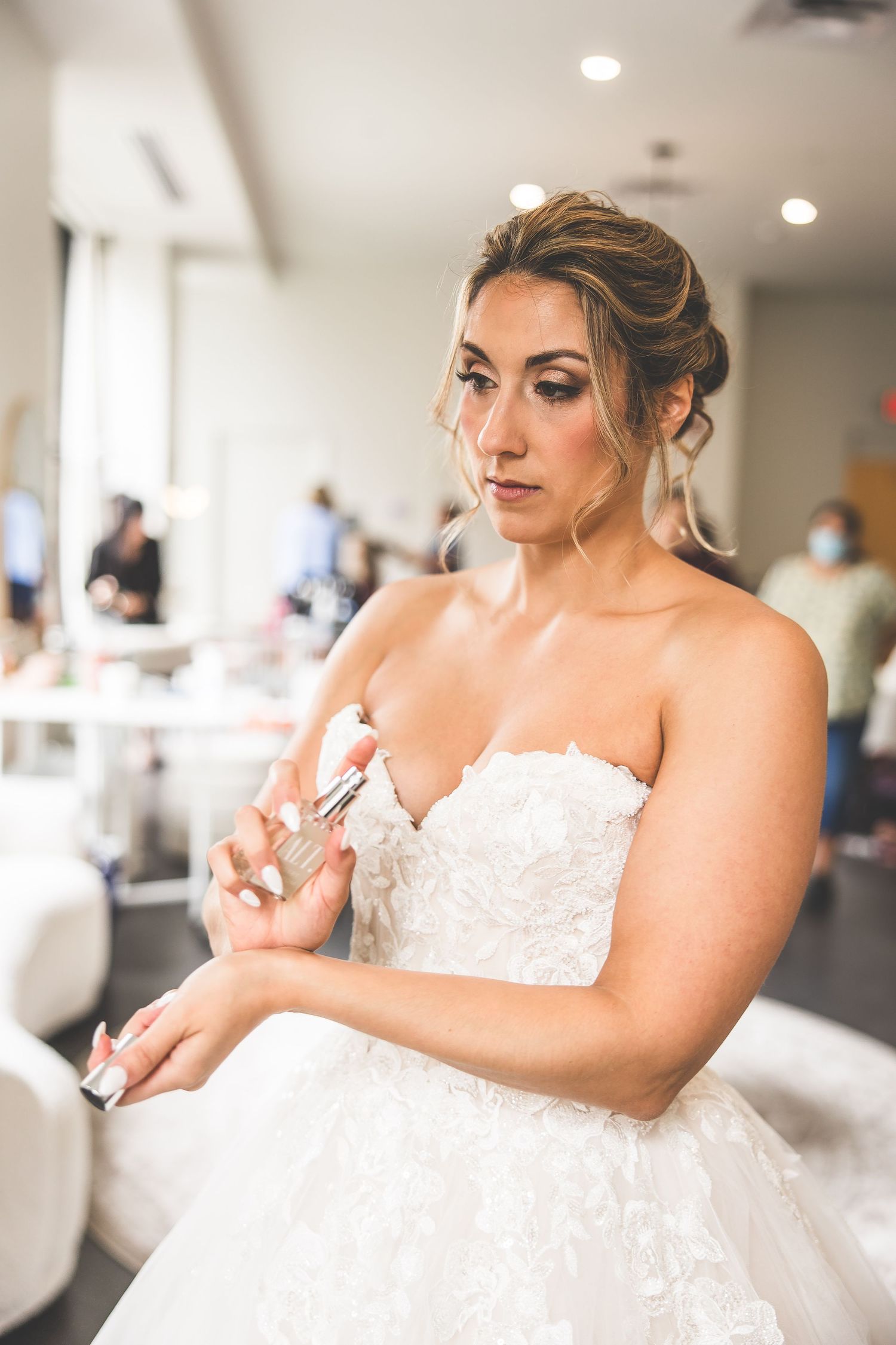 Bride in a strapless lace wedding gown applies perfume while getting ready in a bridal suite.