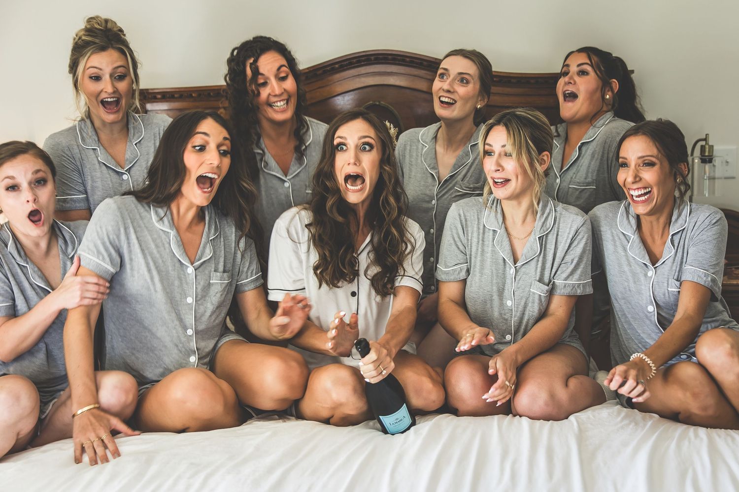 Bridesmaids wearing matching gray robes lounge together on a bed during wedding preparations.