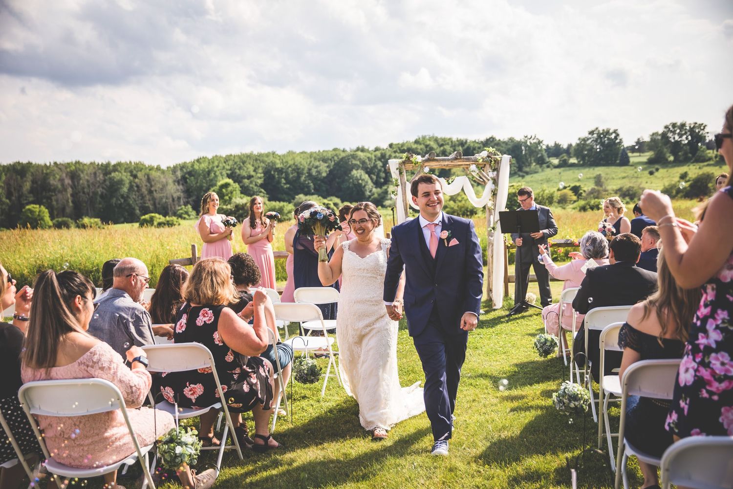 Newlyweds walk down the aisle while guests seated in white chairs celebrate in a sunlit field.