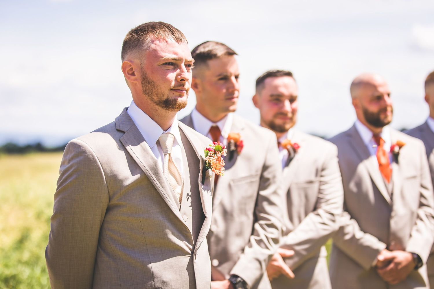 Groomsmen in light gray suits gather outdoors during a summer wedding.