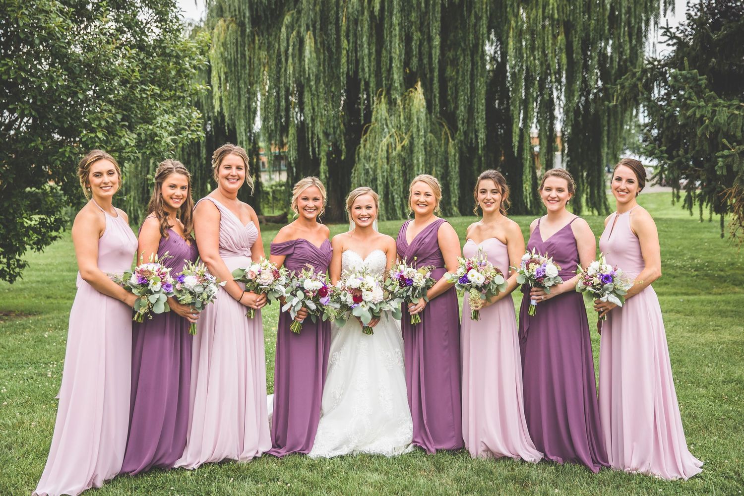 Wedding party in coordinated purple and pink dresses standing together outdoors under willow trees.