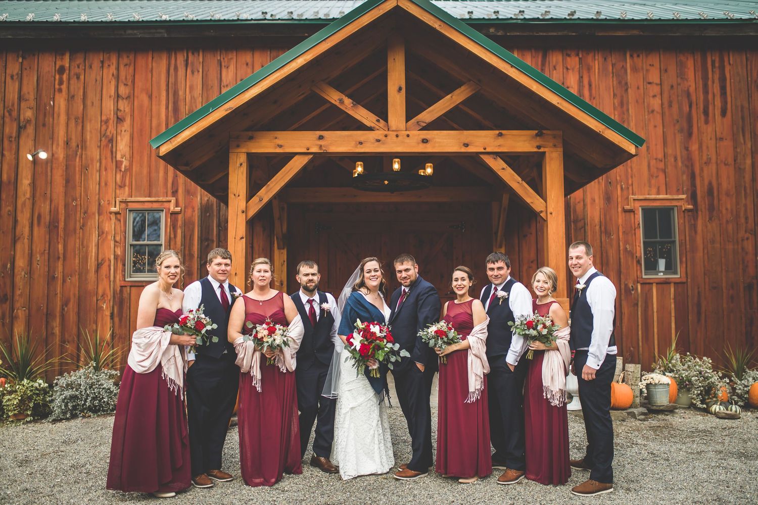 Wedding party in burgundy and black attire poses in front of a rustic wooden barn.