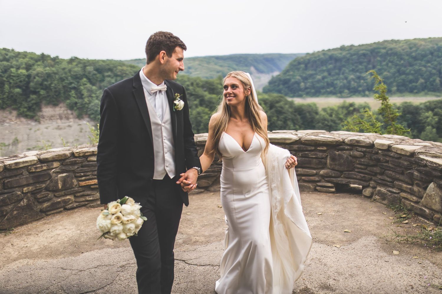 Newlyweds share tender moments along scenic mountain overlook with stone wall.
