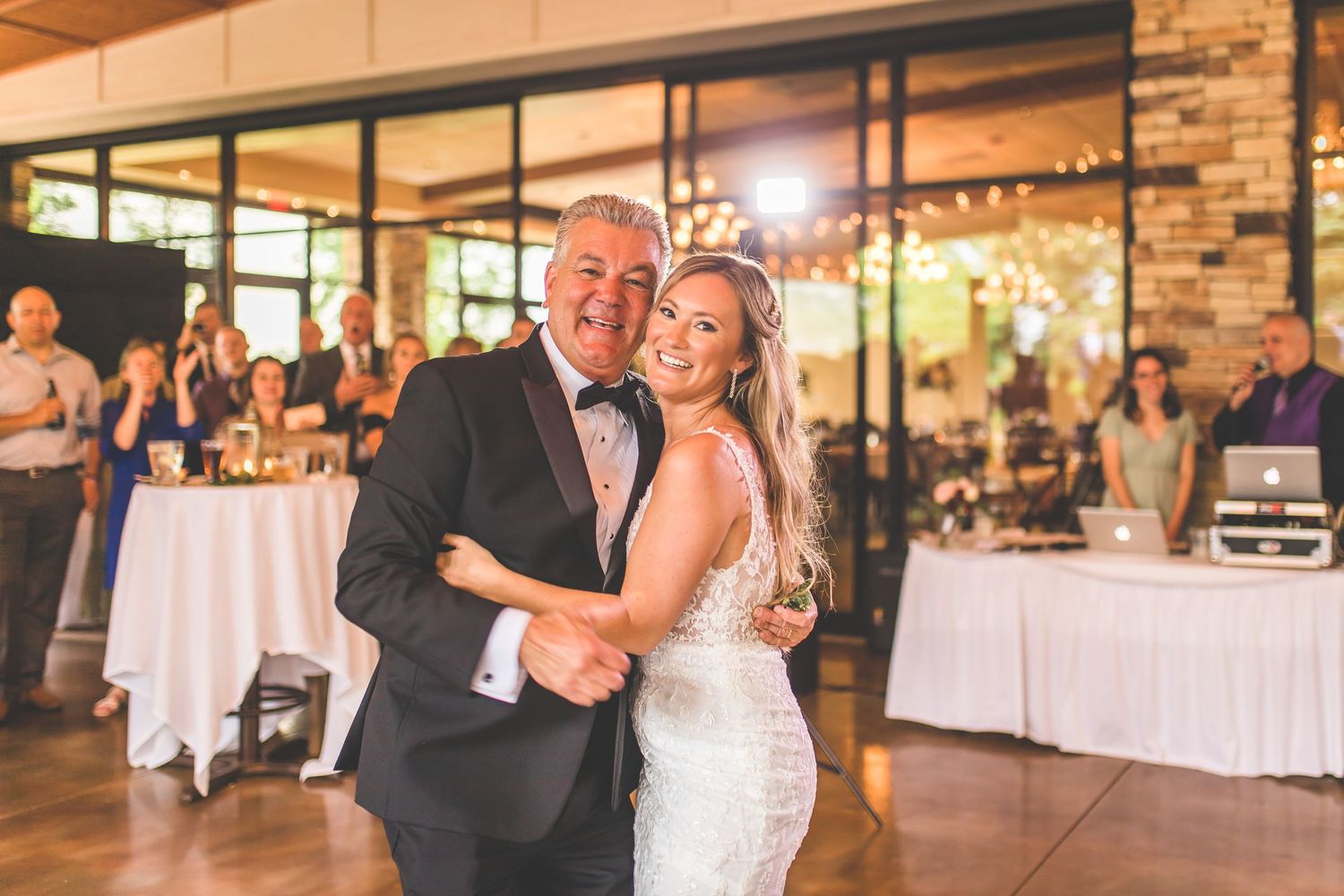 Wedding reception dance moment captured in warm lighting with white tables and stone walls in background.