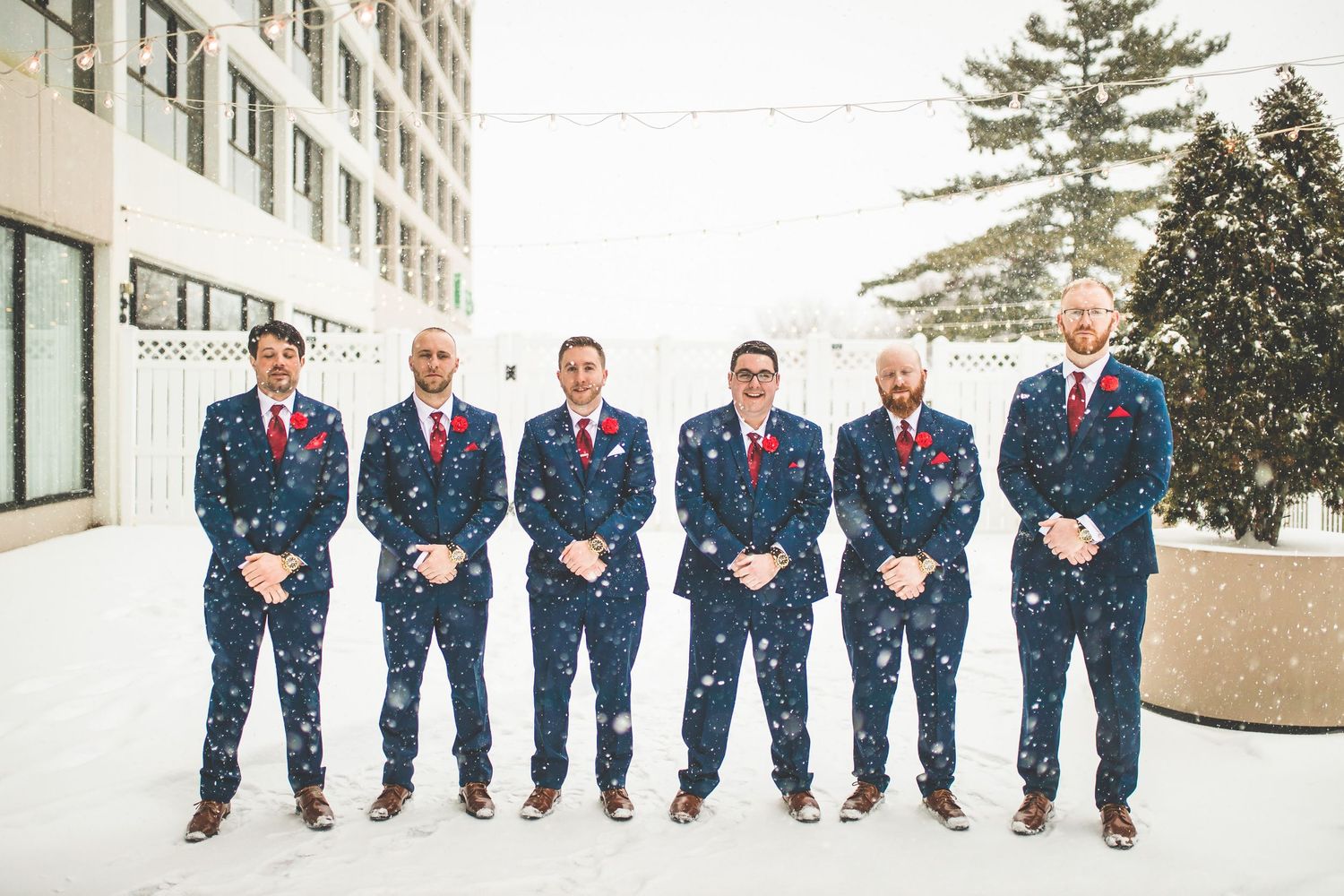 Groomsmen in navy suits with red ties stand in a snowy winter scene outside a building.
