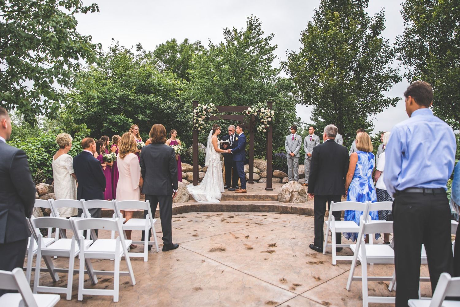 Outdoor wedding ceremony setup with white chairs and floral arrangements under trees.