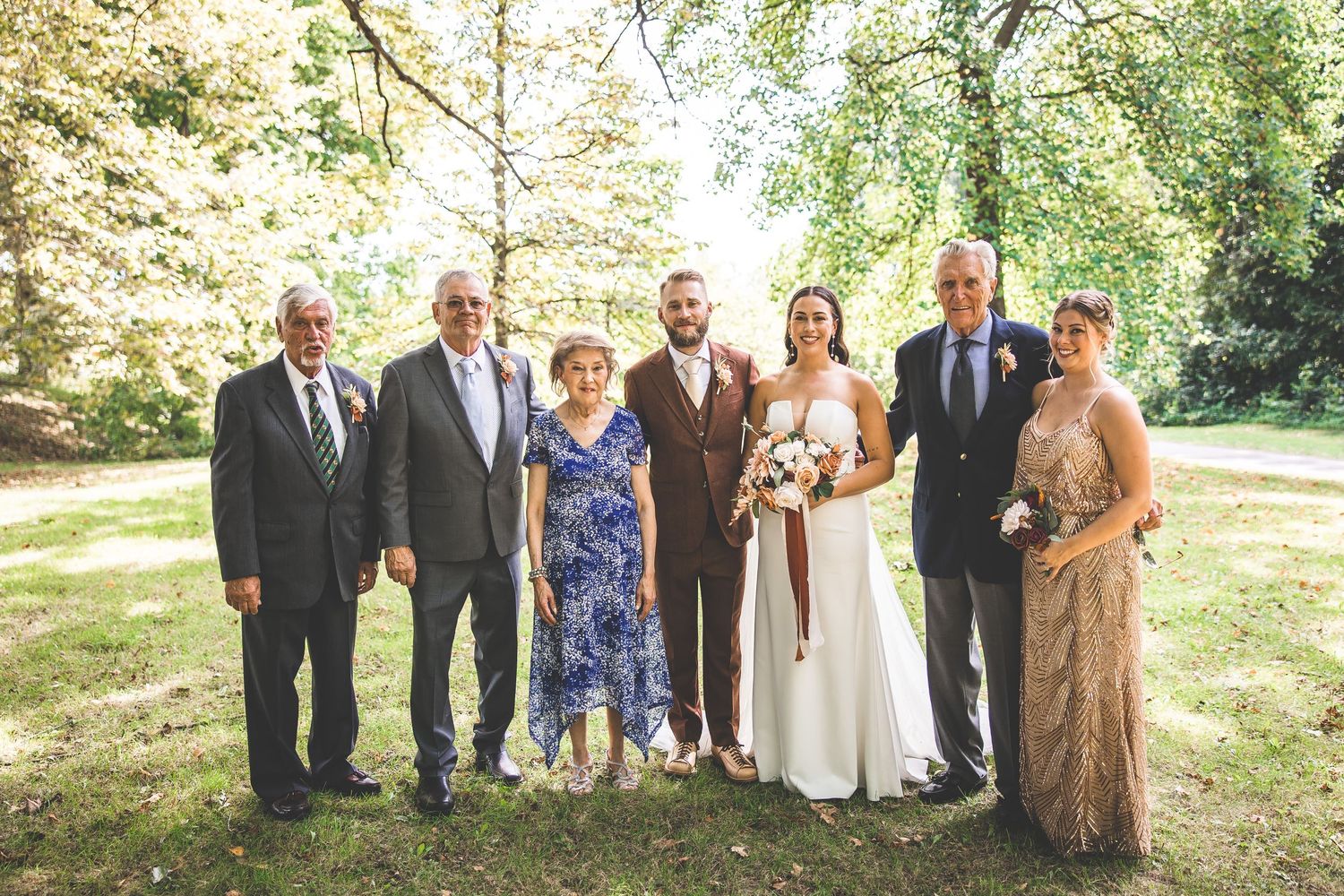 Large wedding group photo with formal attire arranged under trees in afternoon light.