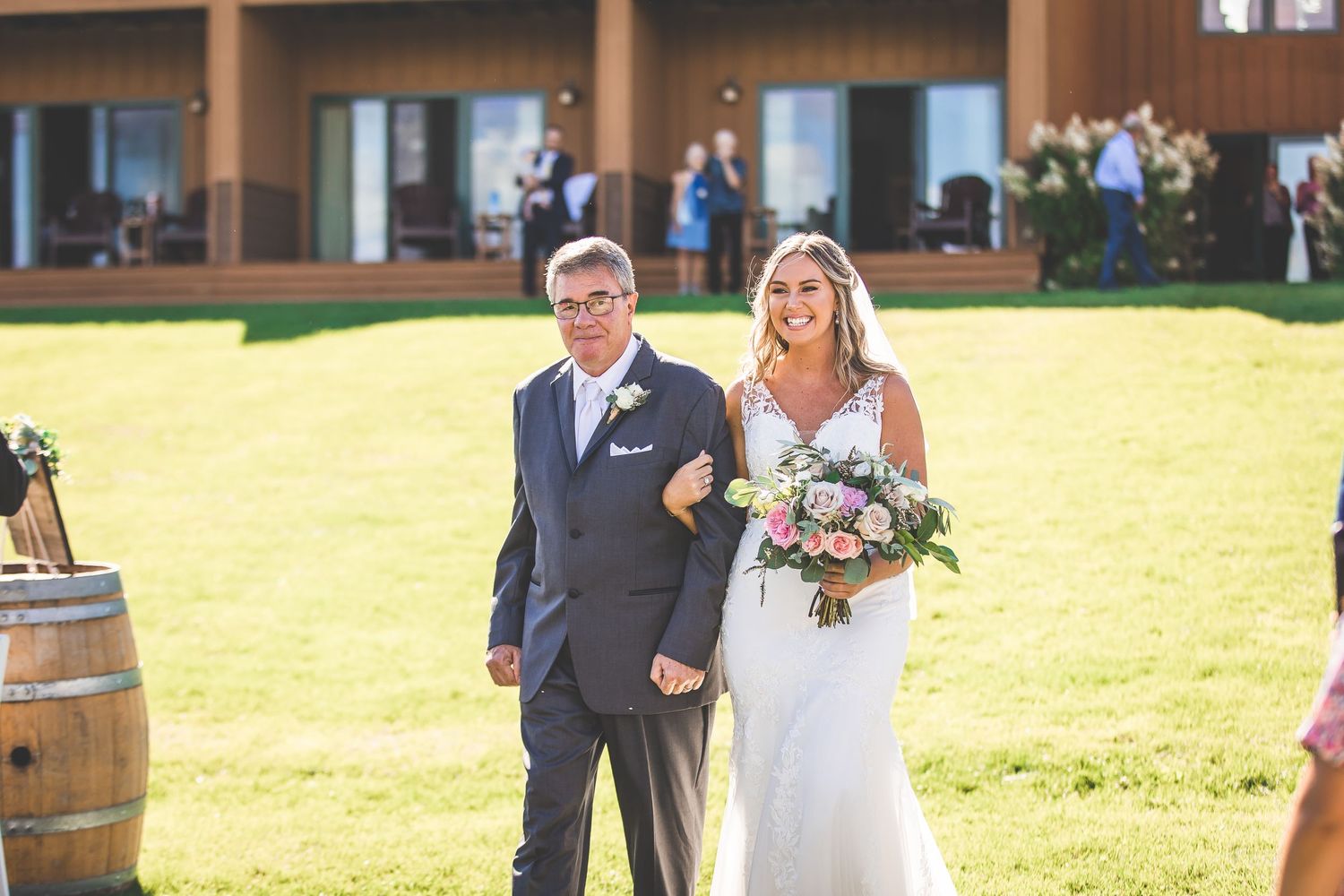 A wedding ceremony takes place on a sunny lawn with wine barrels and guests gathered in the background.