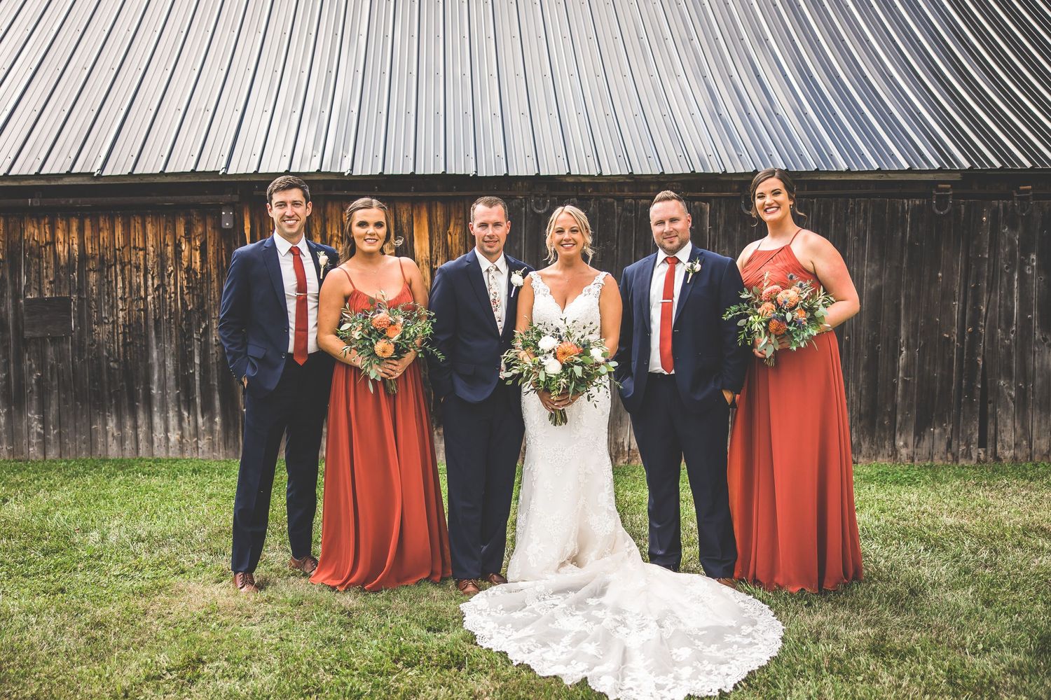 Wedding party in navy suits and rust-colored dresses pose together in front of rustic barn backdrop.