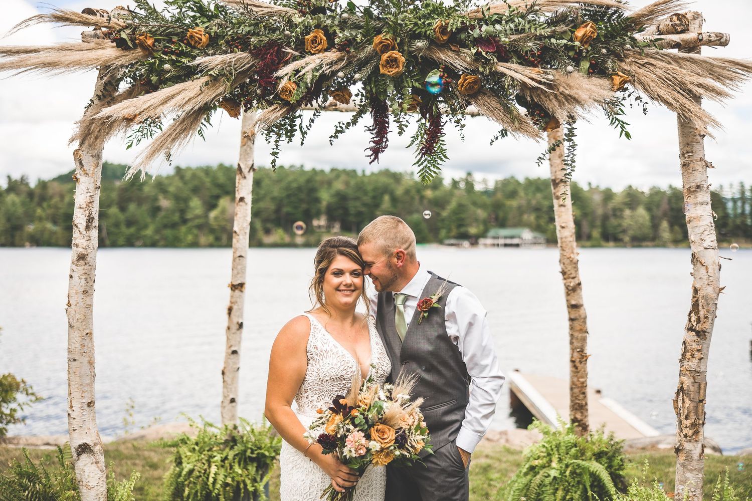Rustic birch branch wedding arch decorated with greenery and flowers overlooking a lake.