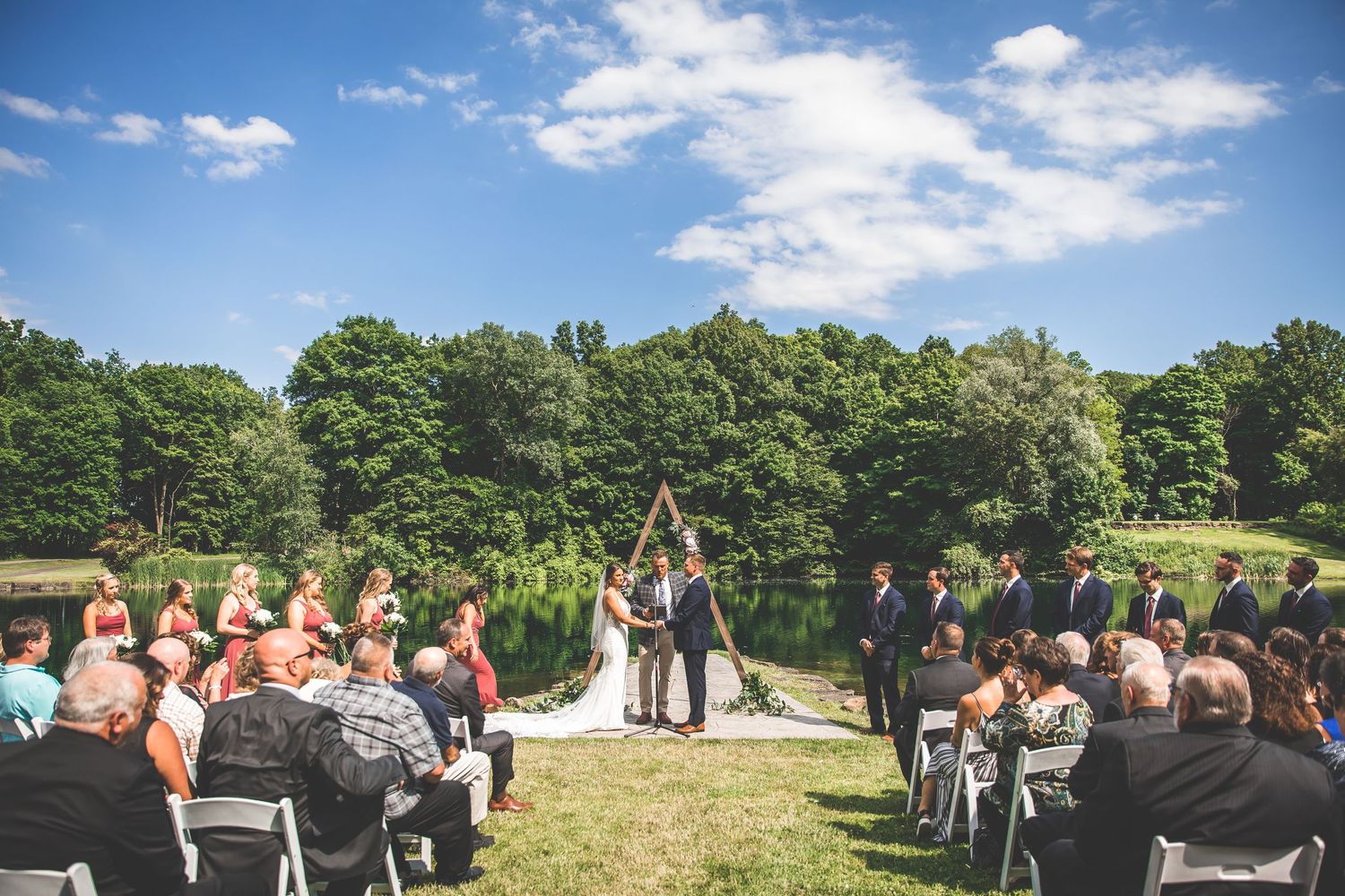 Outdoor wedding ceremony setup with white chairs and guests gathering on a sunny summer day by a lake.