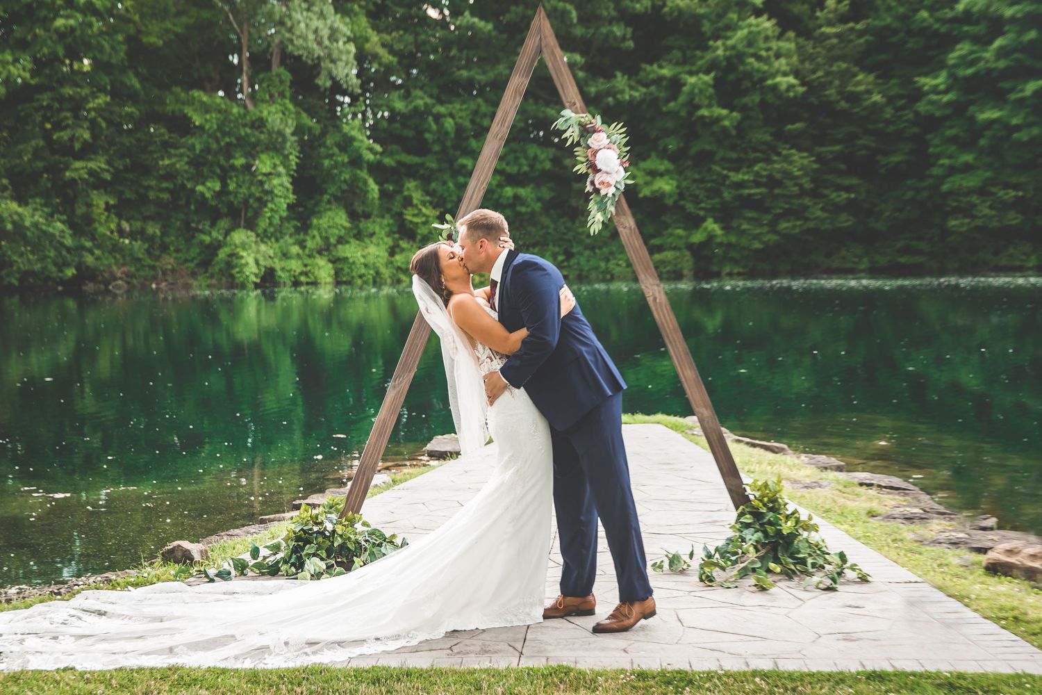 Wedding ceremony in progress by lakeside with wooden triangle arch and natural setting.