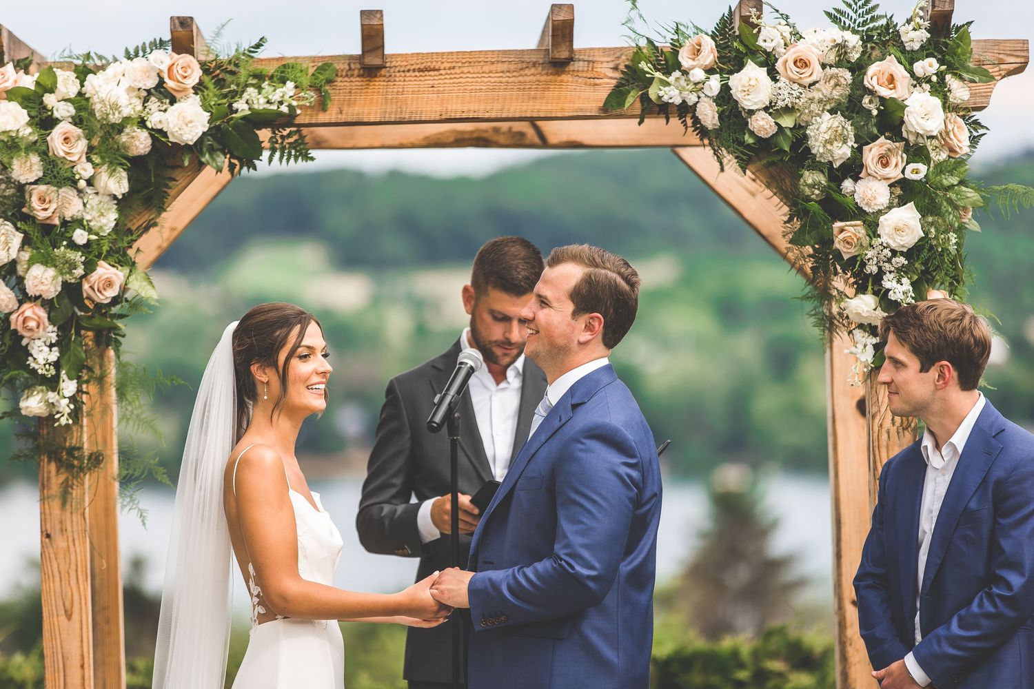 Wooden wedding arch decorated with white roses and greenery against a scenic landscape backdrop.