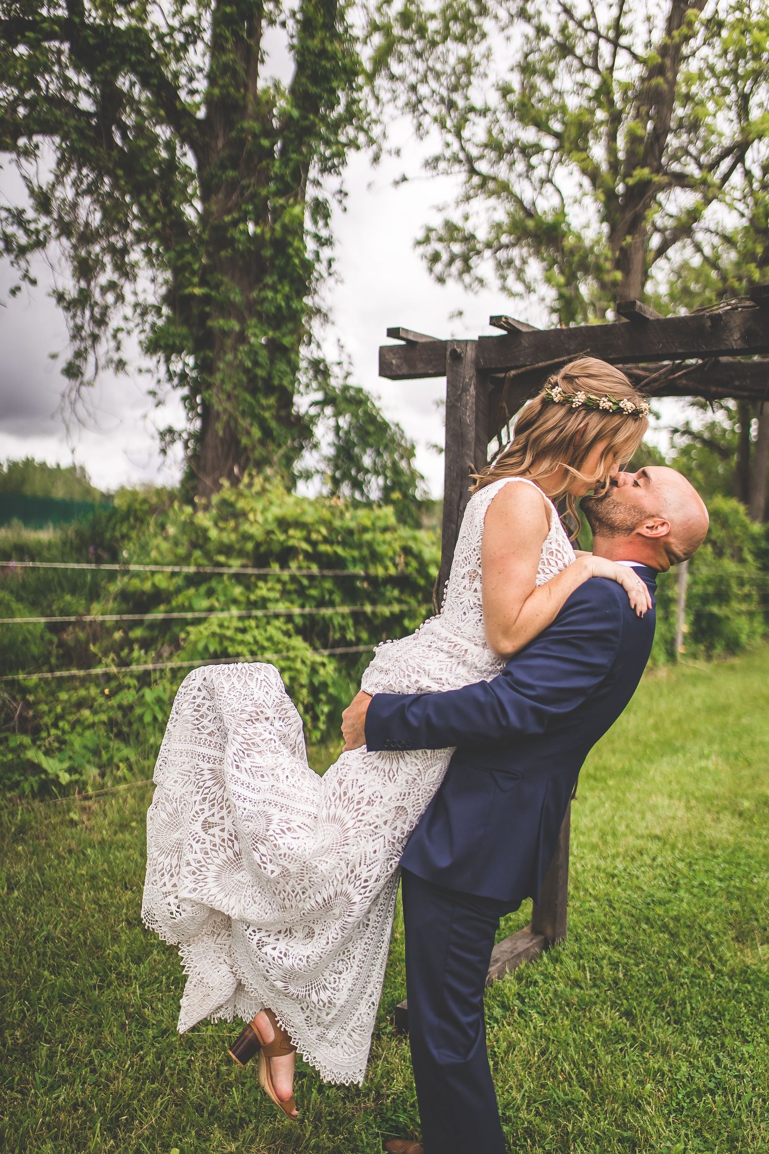A joyful wedding lift pose beneath a wooden garden arbor with trailing lace dress.