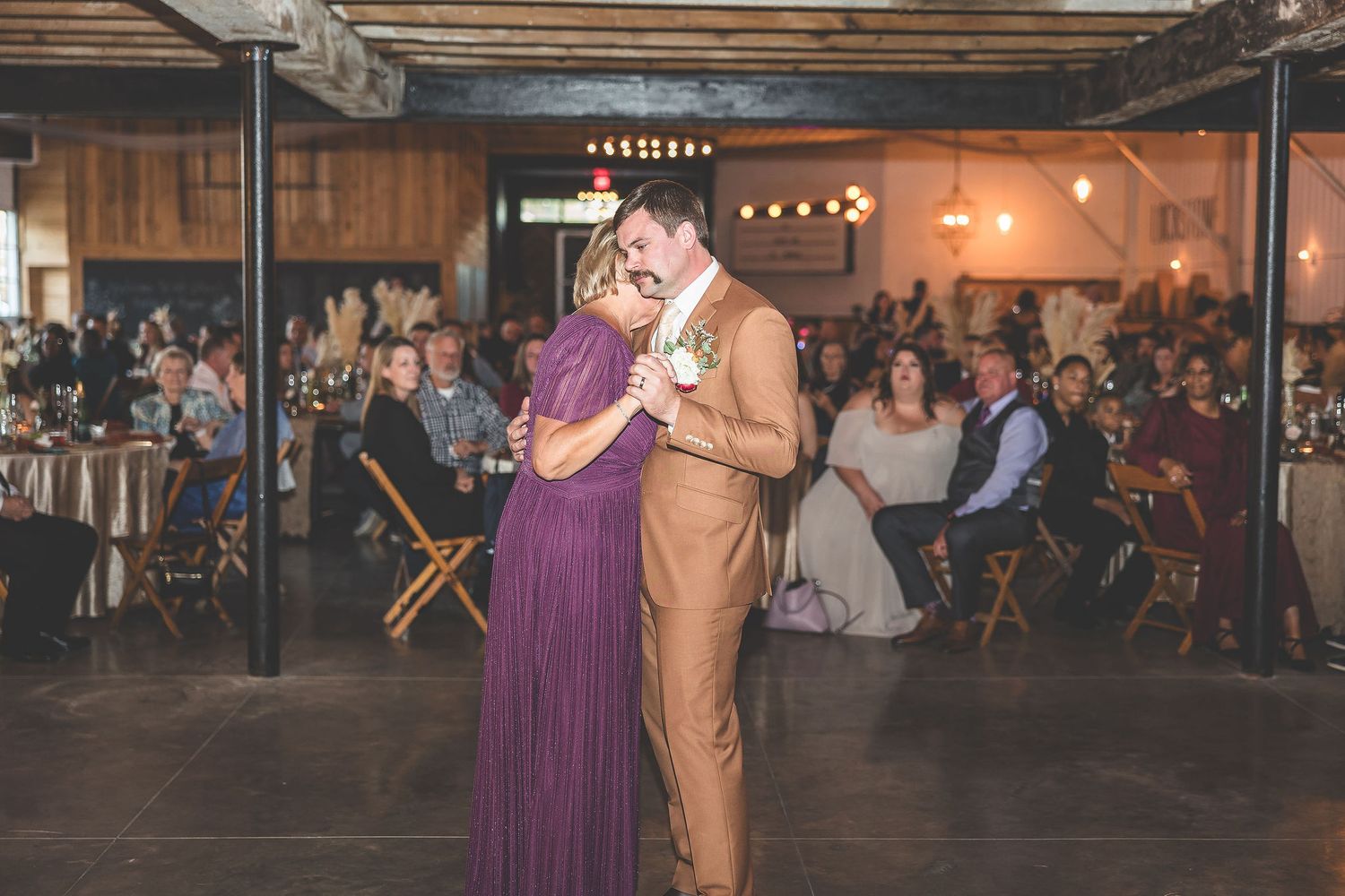 Wedding reception dance floor filled with couples dancing under string lights in rustic venue.