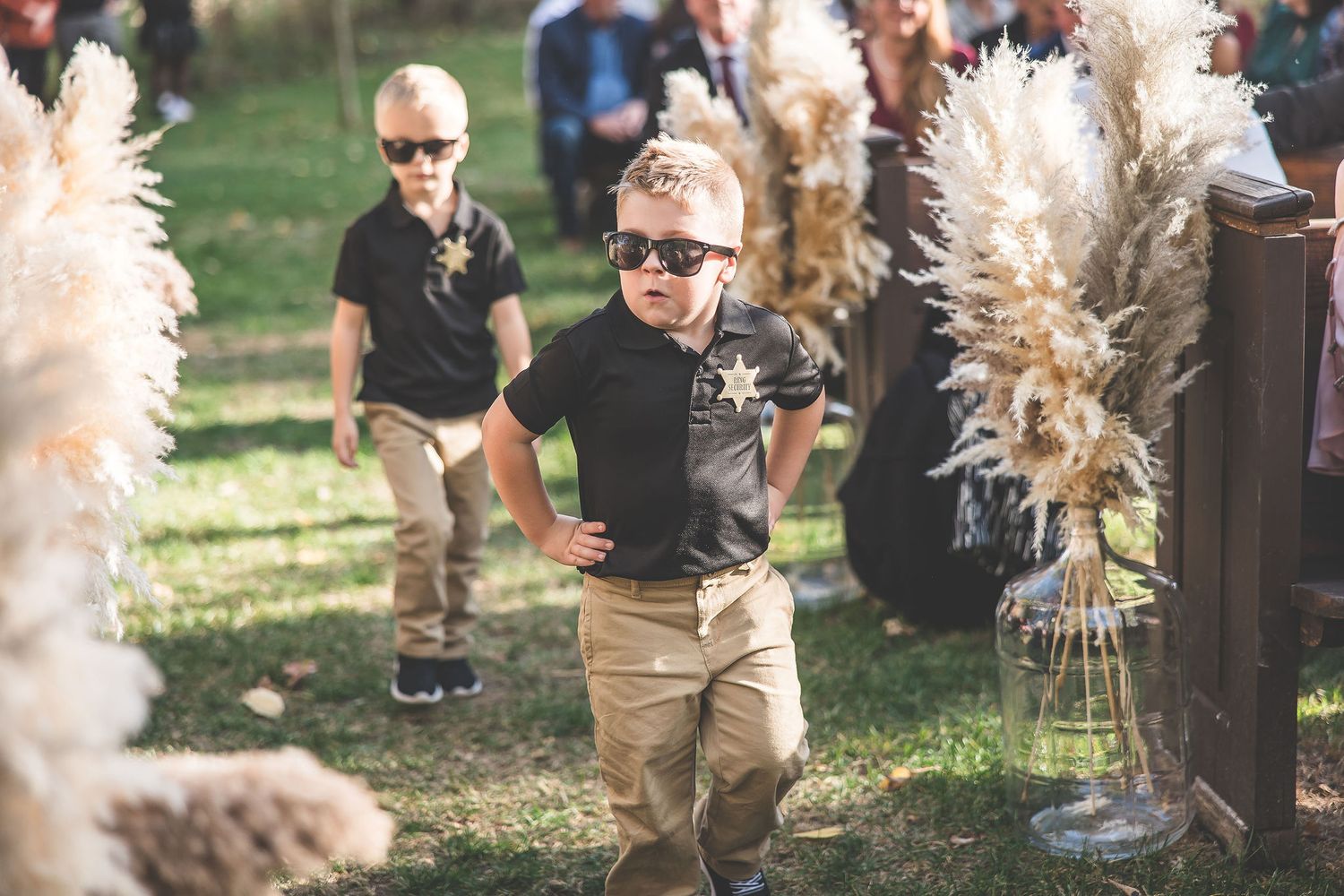 Young ring bearers in black polos and khaki pants walk between pampas grass decorations.