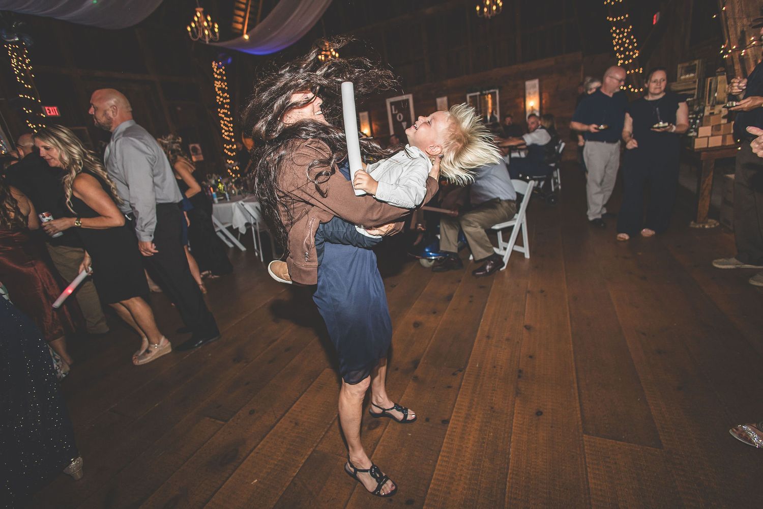 Wedding guests dance and celebrate on a rustic wooden dance floor at an indoor reception venue.