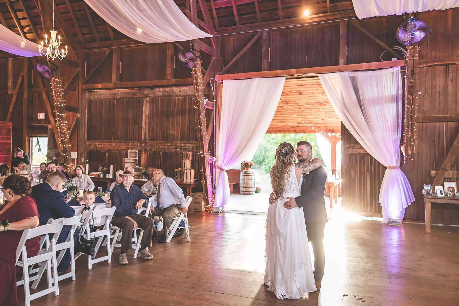 Rustic barn wedding reception with elegant white draping and purple accent lighting creating romantic atmosphere.