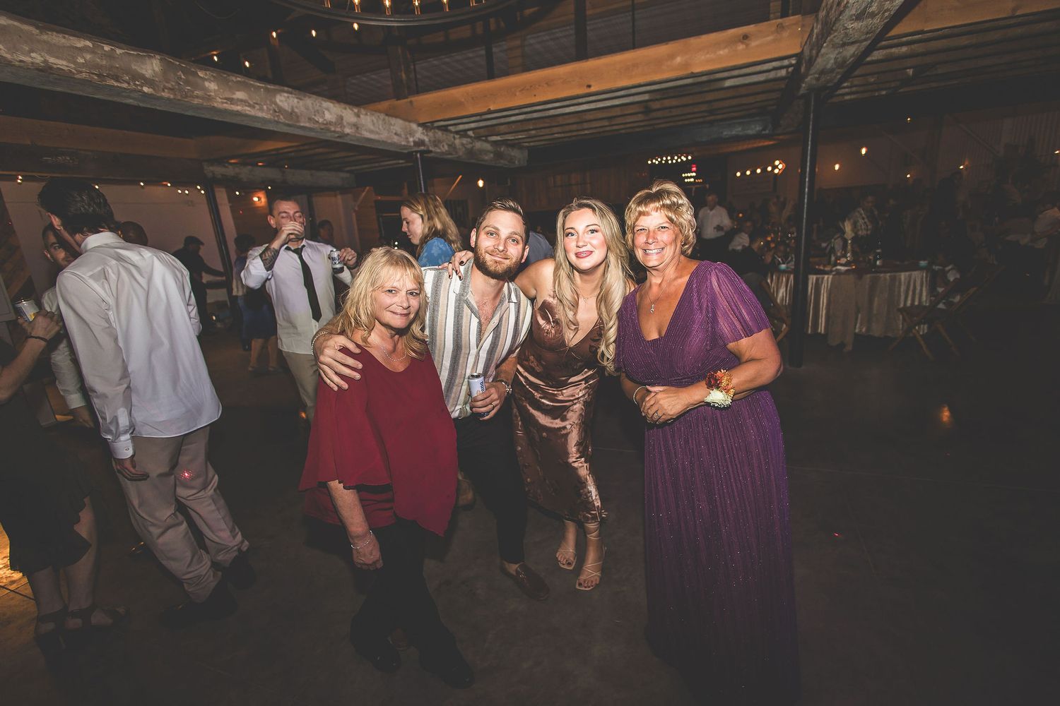 Wedding reception guests dance and celebrate in a dimly lit rustic barn venue.