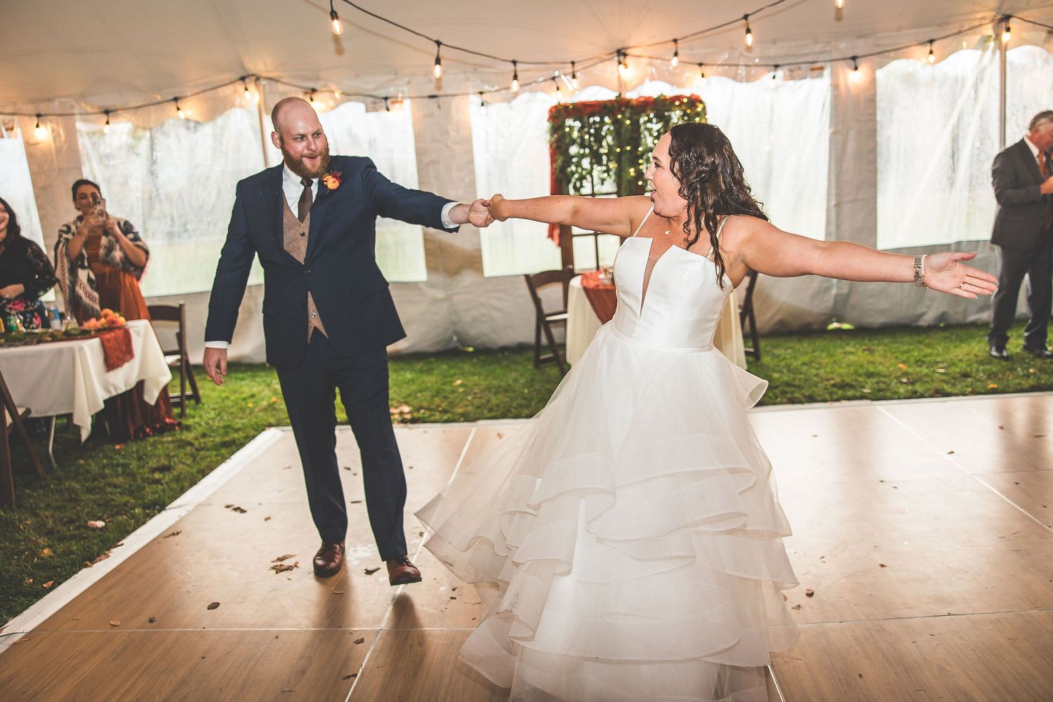 Wedding couple dances under string lights in white tent during reception celebration.