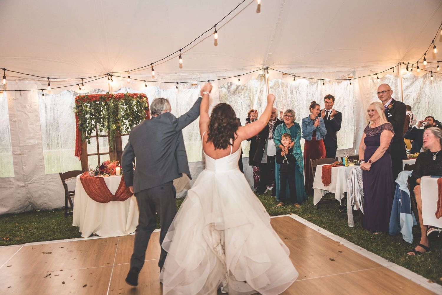 Wedding reception guests dancing and celebrating under string lights in a white tent venue.