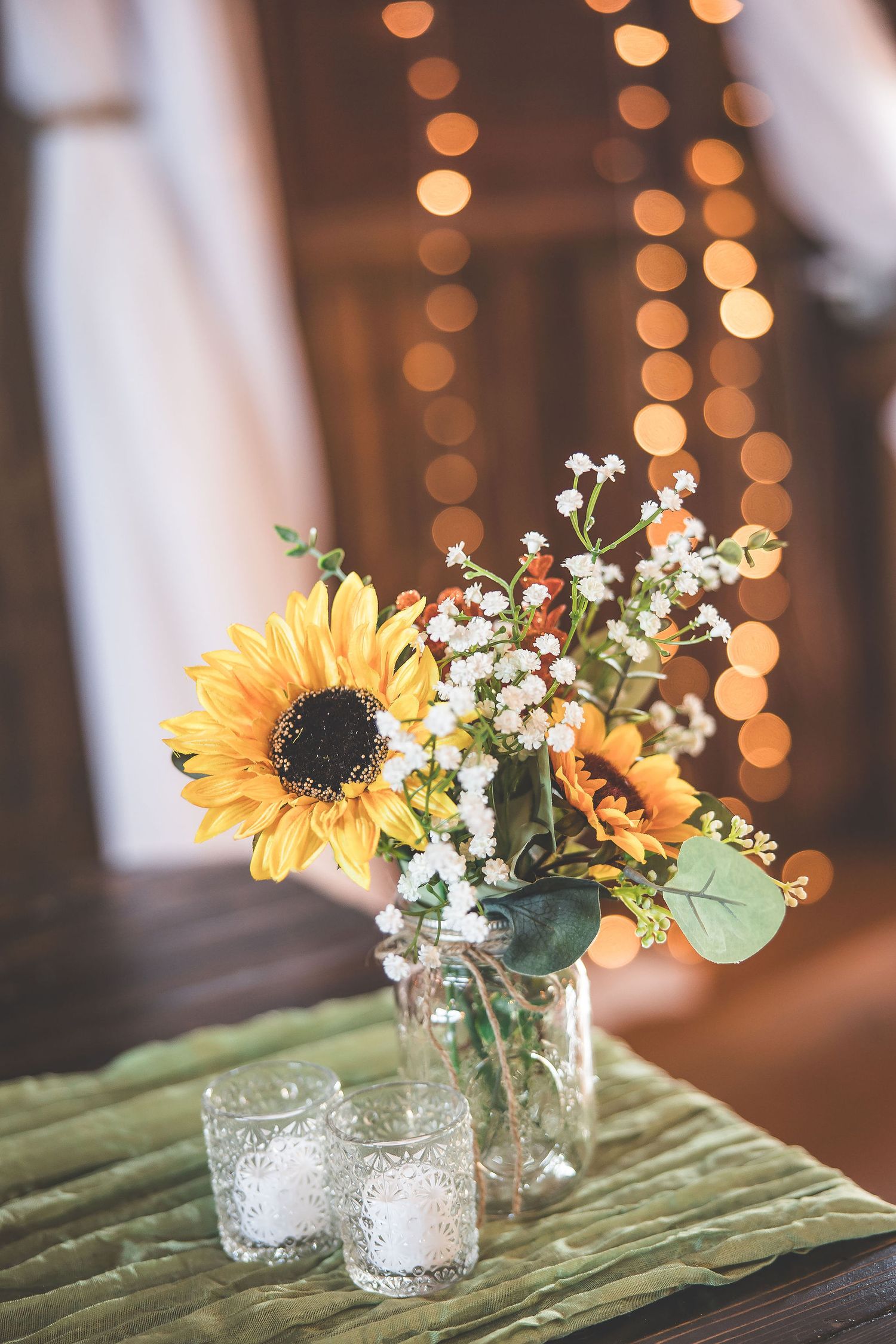 Mason jars with sunflowers and baby's breath centerpieces on wooden table with bokeh lights in background.