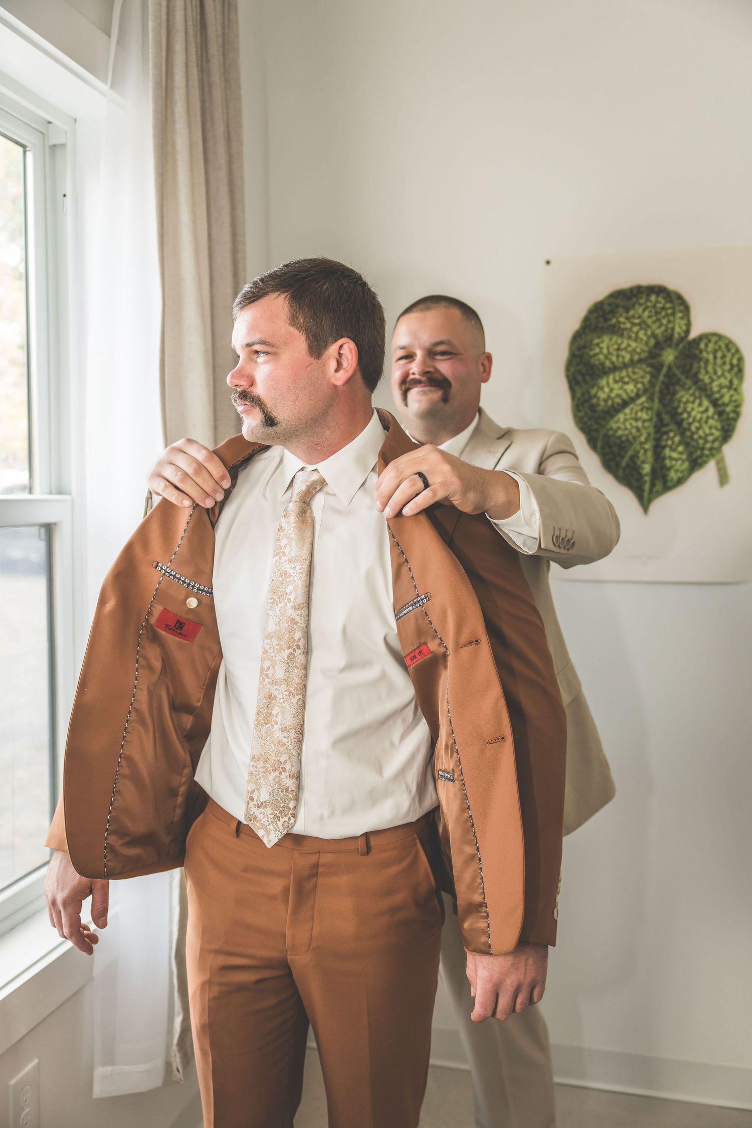 Groomsmen in brown suits pose playfully against a white wall with green leaf decorations.