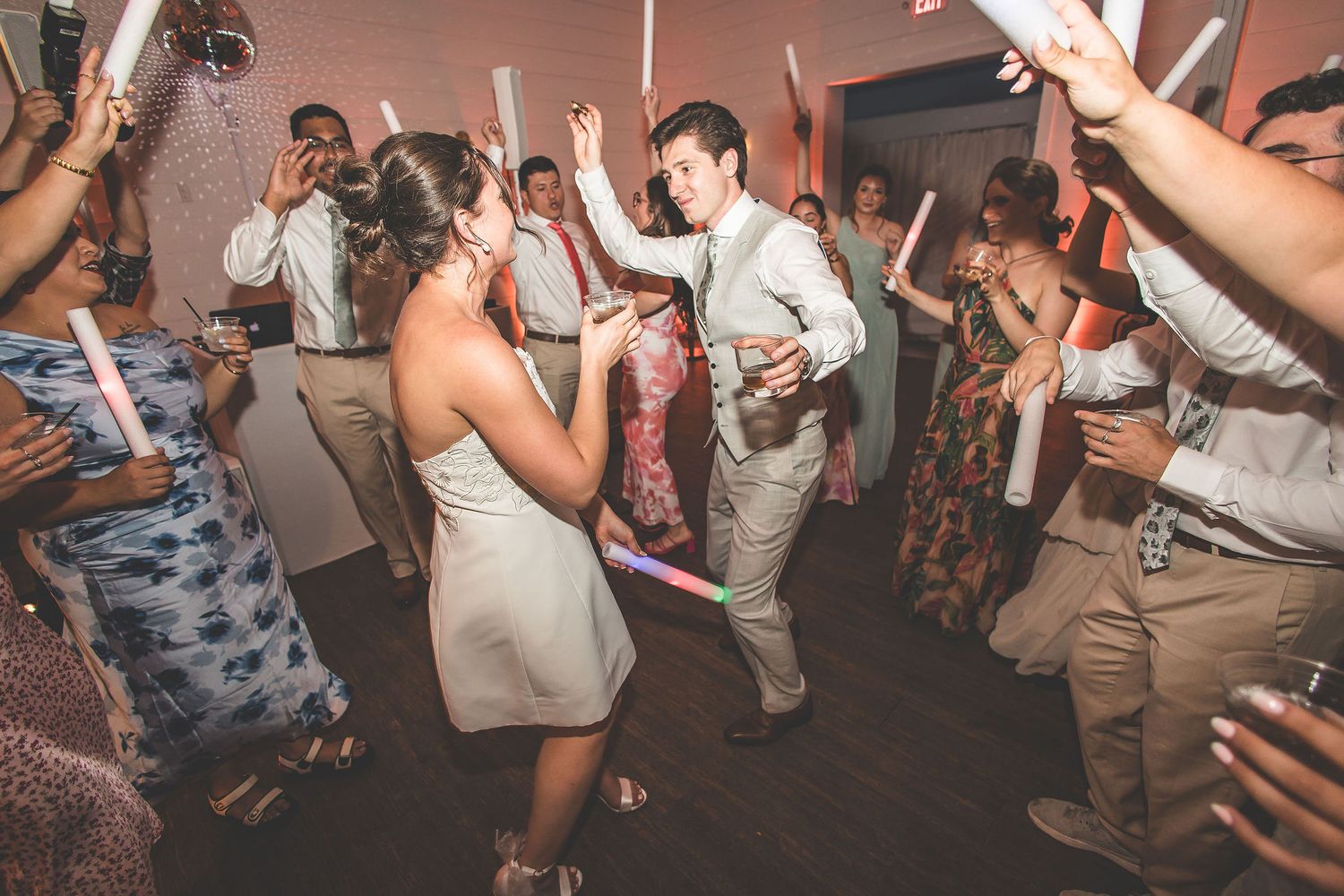 Wedding guests dancing in a circle holding glowing foam sticks at an indoor reception.