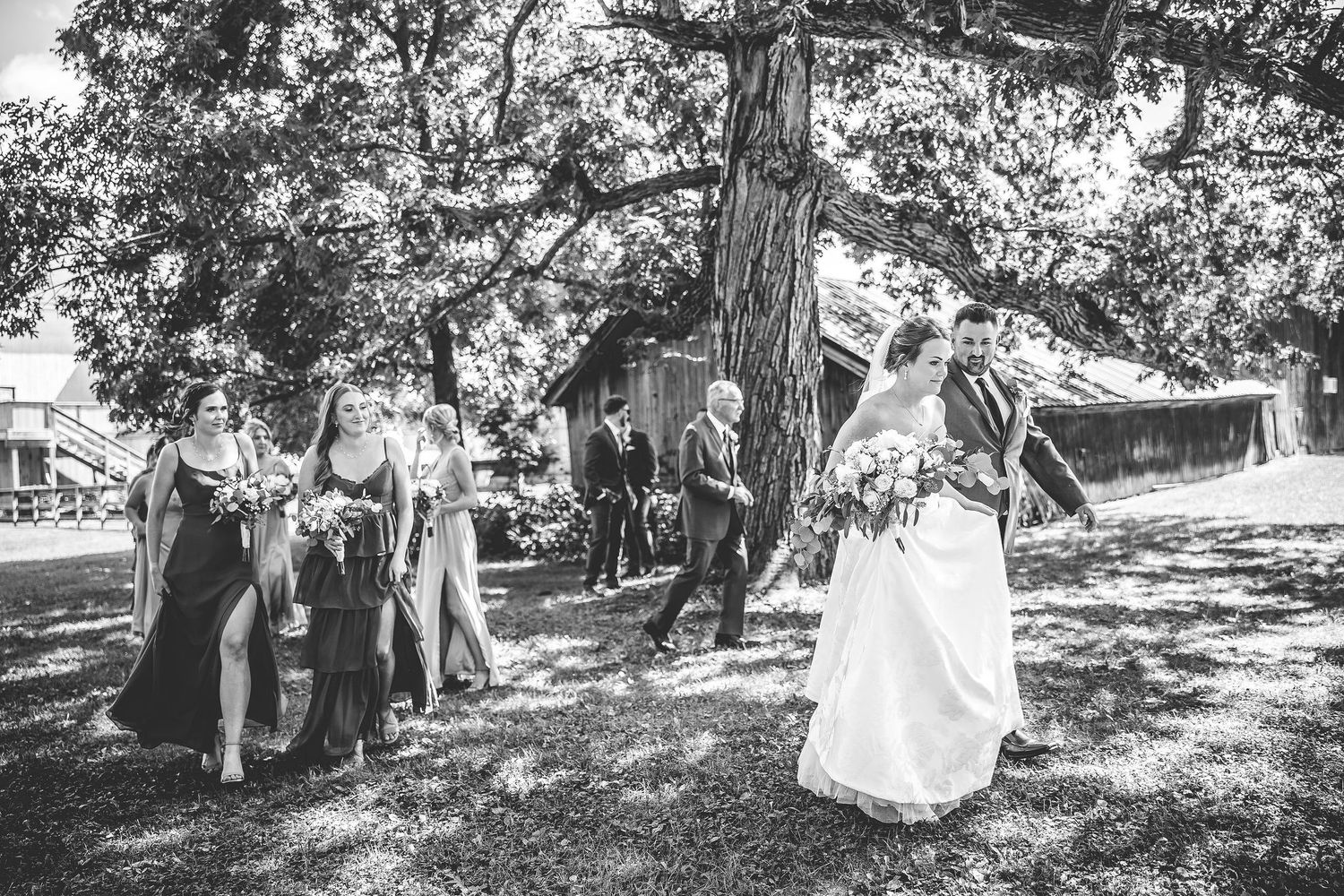 Black and white photo of wedding party members posing outdoors under large trees.