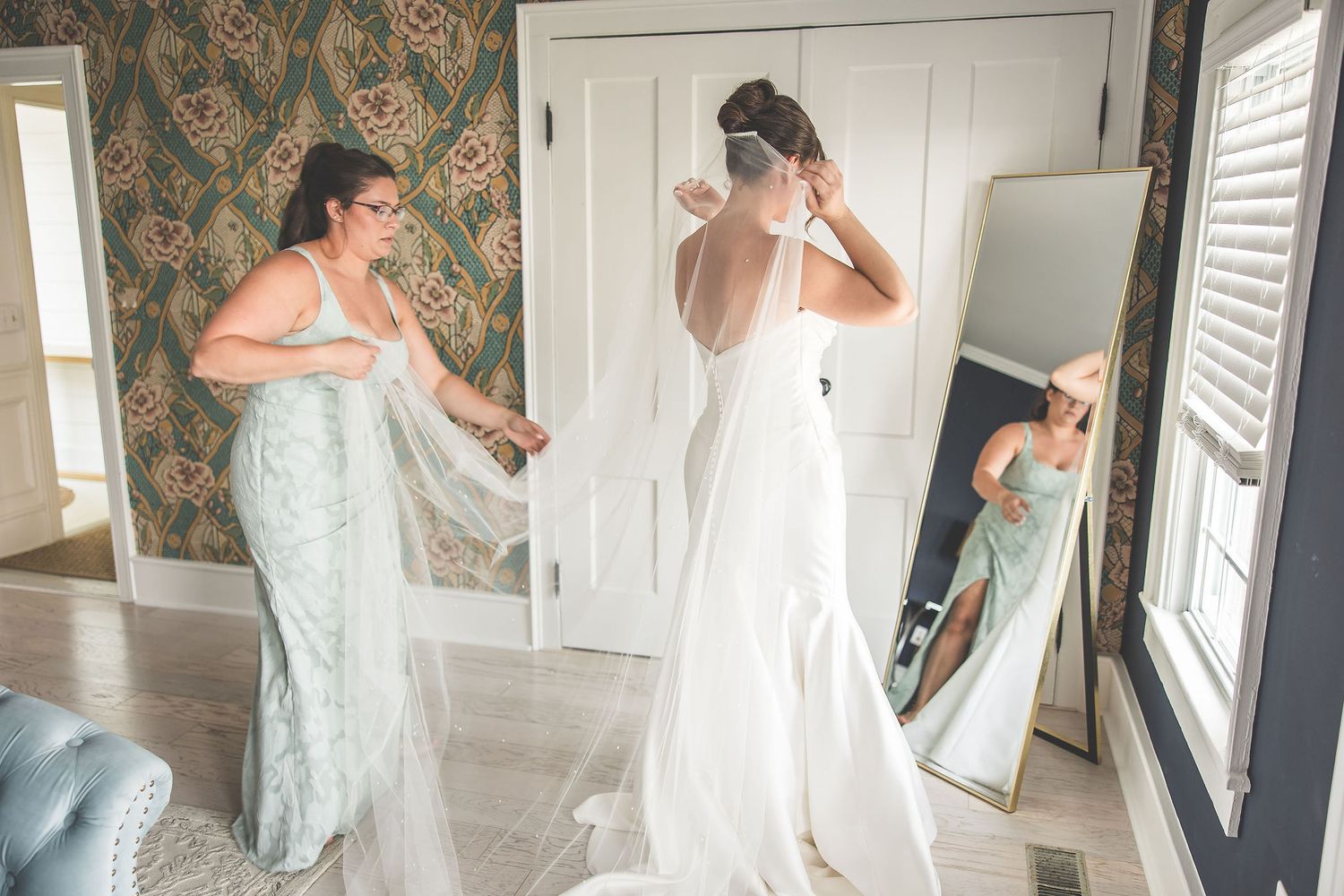 Two people getting ready in formal attire with floral wallpaper and mirrors in the background.