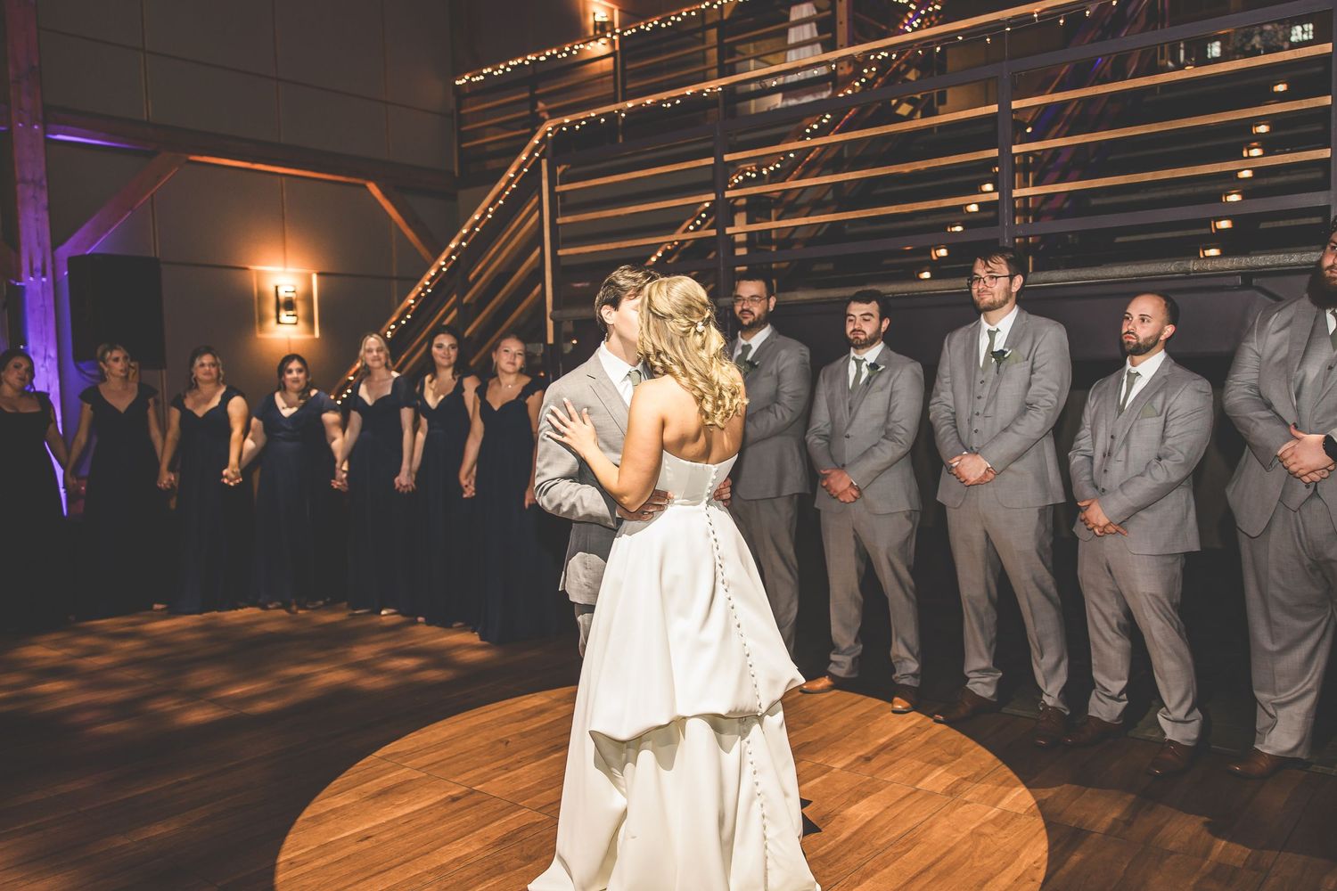 A romantic first dance moment captured on a circular wooden dance floor with wedding party looking on.