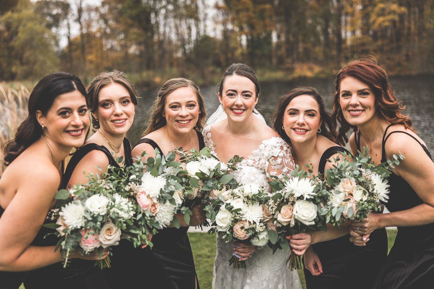 Bridesmaids in black dresses hold white bouquets during autumn wedding celebration.
