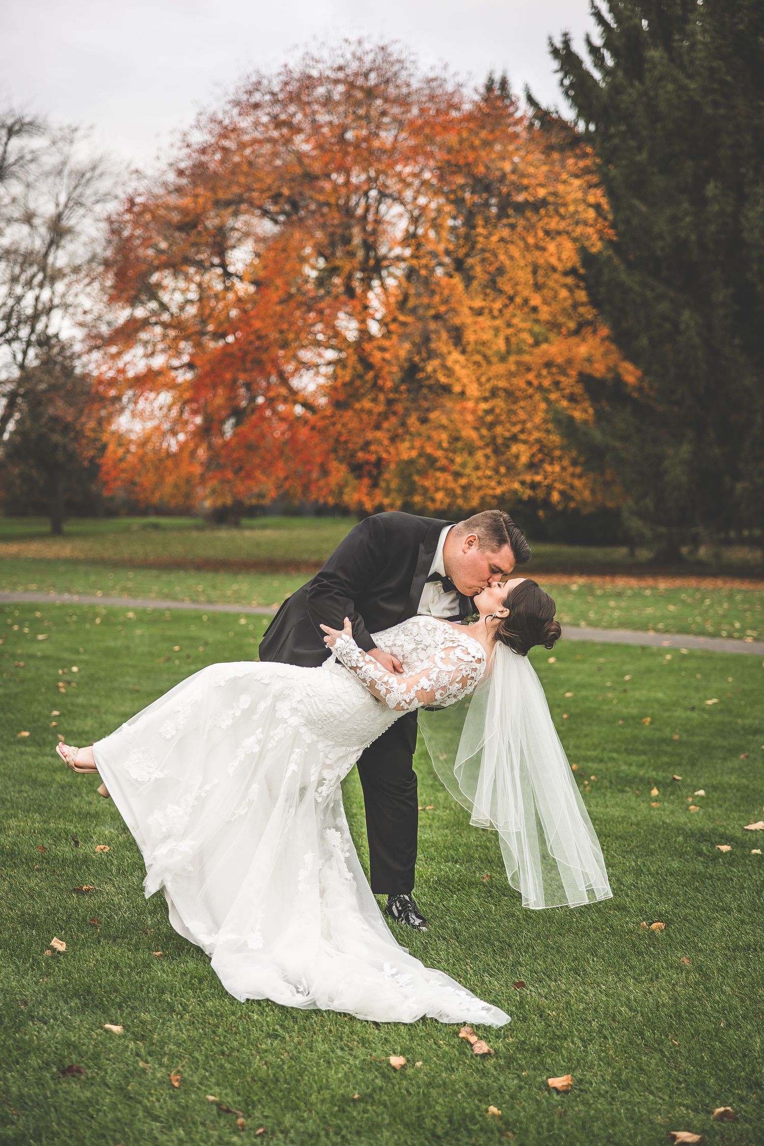 A romantic wedding couple dips and spins on a grassy lawn surrounded by vibrant autumn foliage.