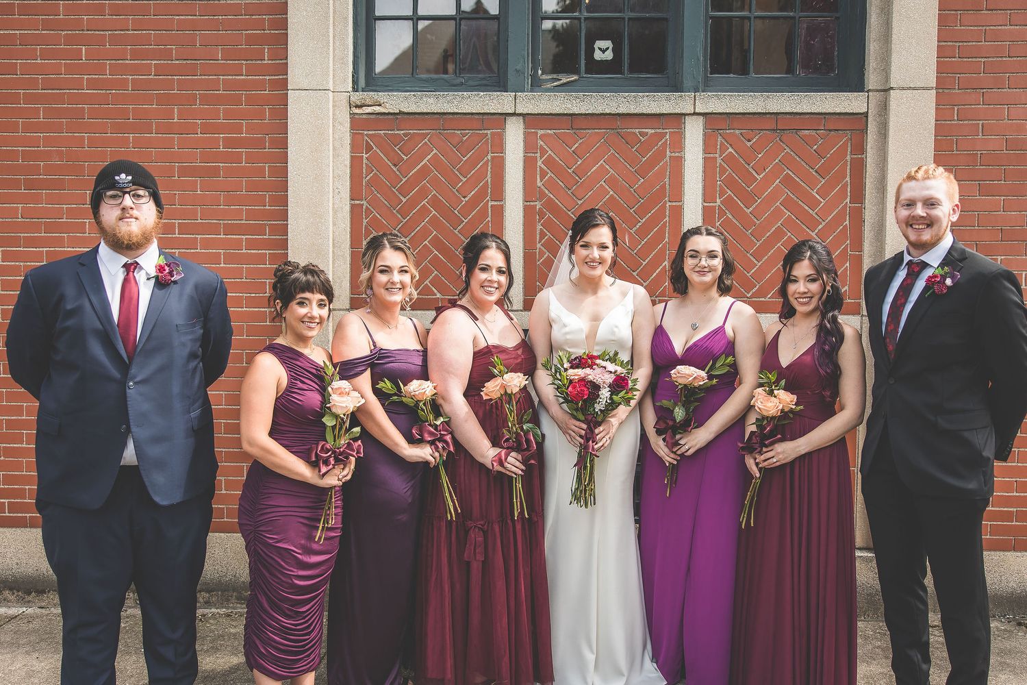 Wedding party in burgundy and navy attire lined up against brick building exterior.