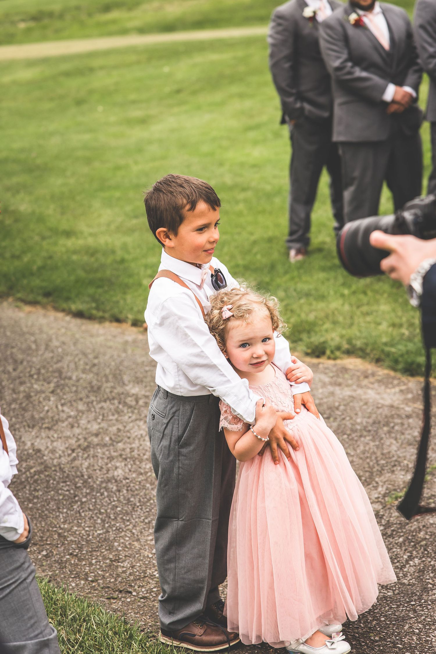 Two young children in formal attire share a sweet embrace at an outdoor wedding celebration.