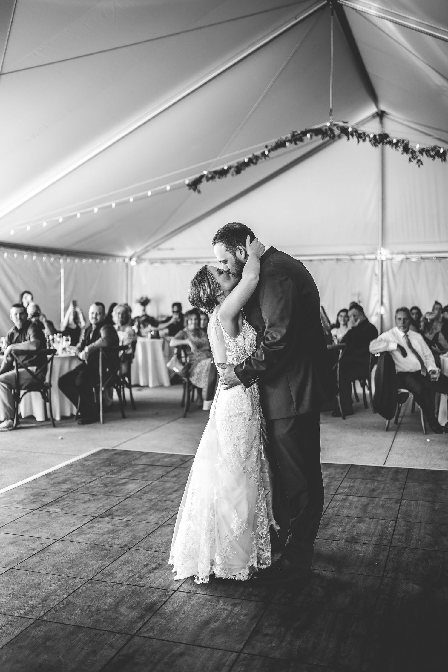 Black and white photo of a wedding couple's first dance surrounded by guests in a tented venue.