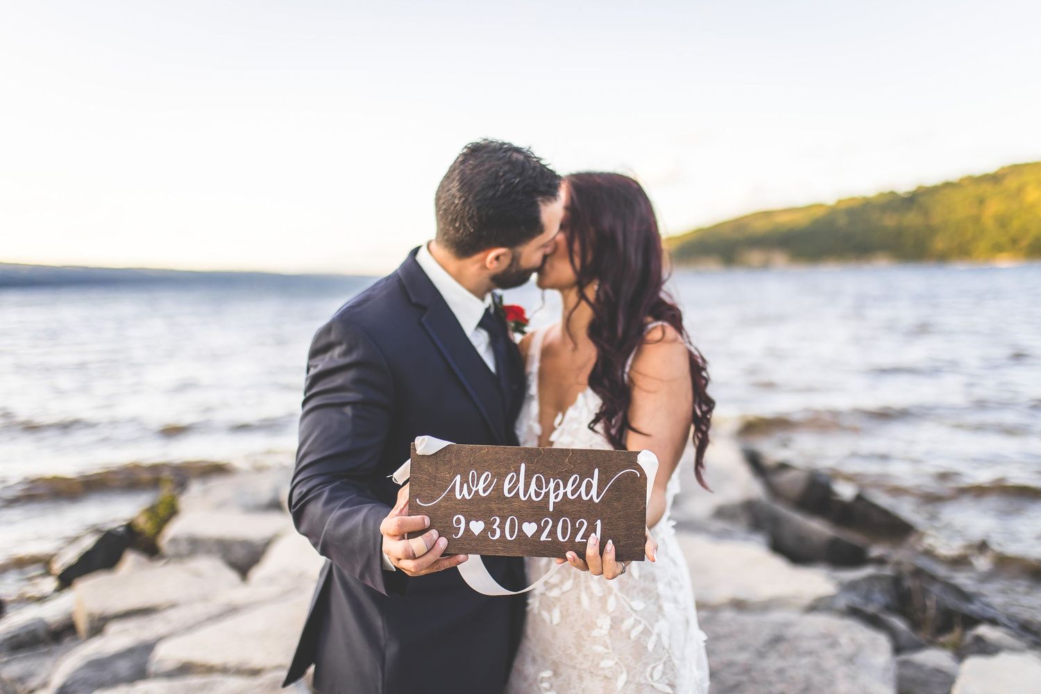 Newlyweds pose with wooden sign on rocky beach at sunset.