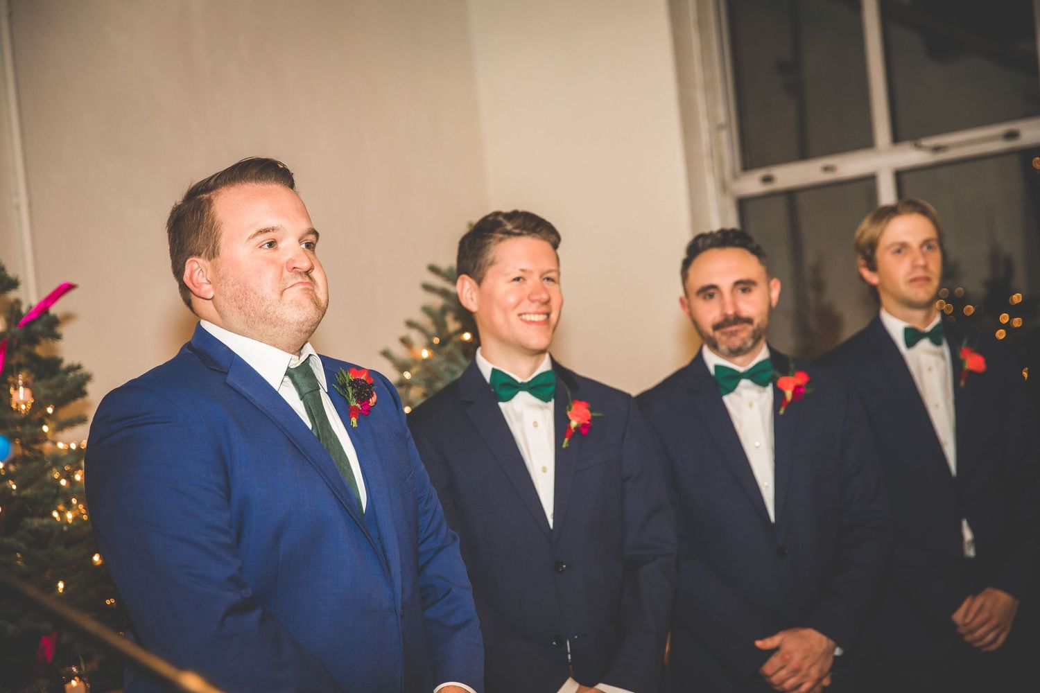 Groomsmen in navy suits stand together in front of Christmas decorations at an indoor venue.