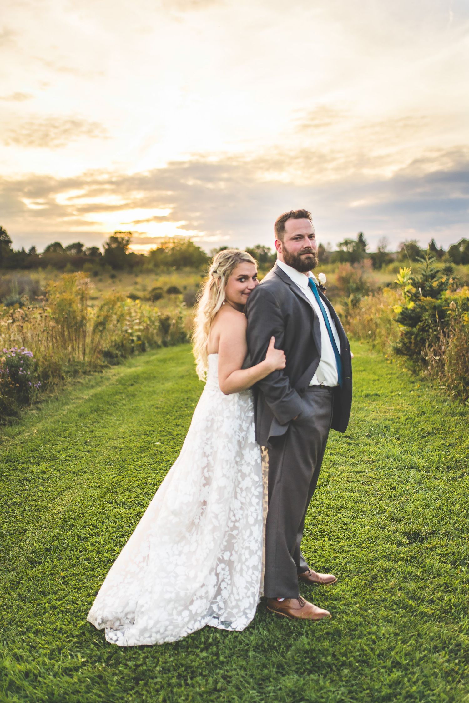 A couple in wedding attire poses together on a grassy hillside during golden hour sunset.