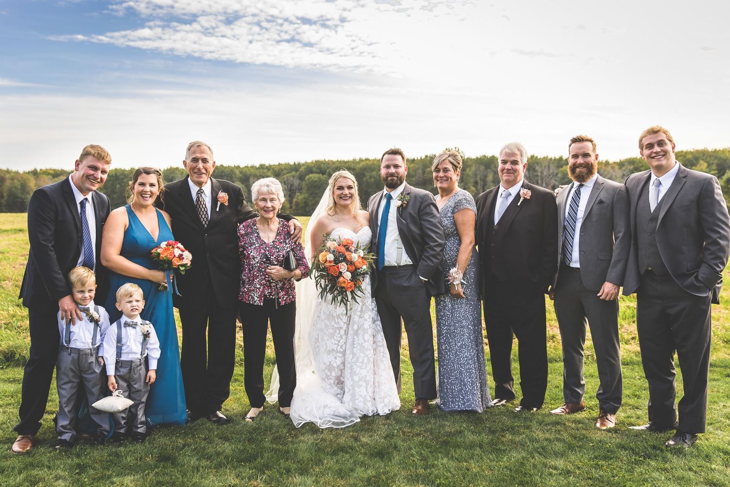 Large wedding party group photo on grassy hillside under blue sky.