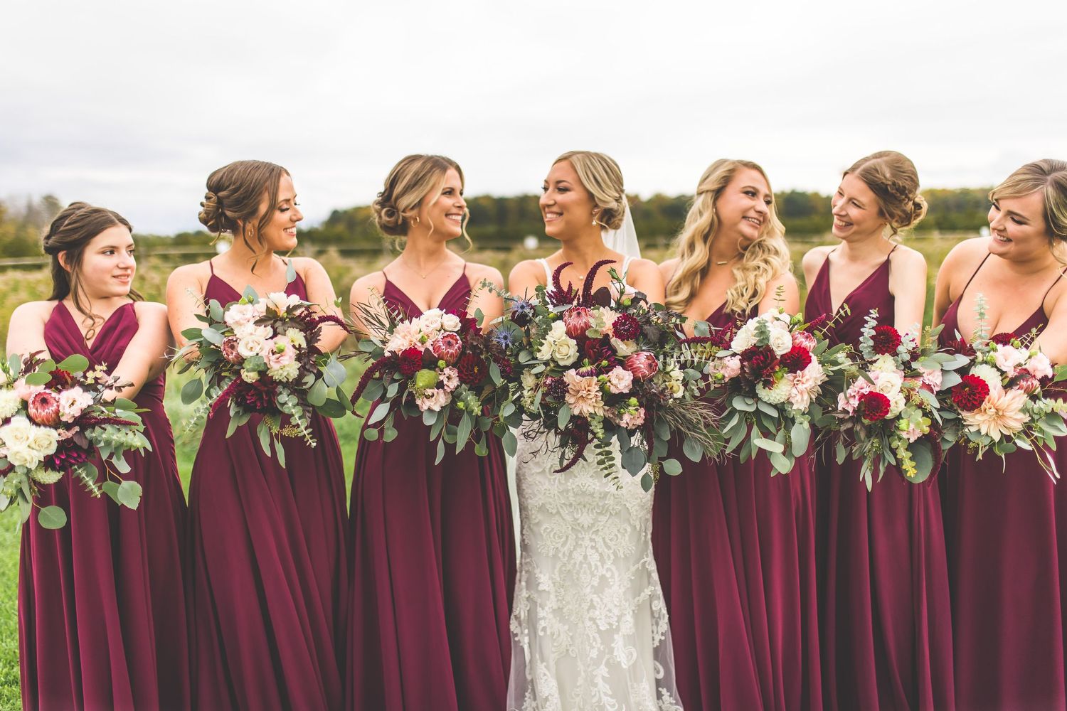 Bridesmaids in burgundy dresses laugh together during outdoor wedding photos.
