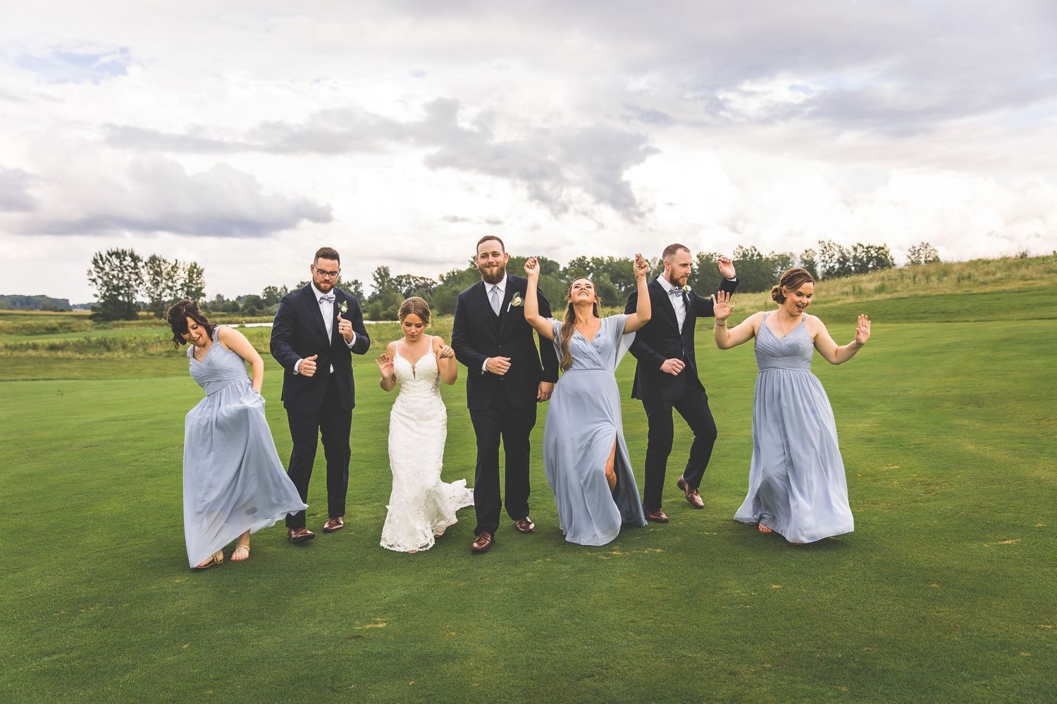 Wedding party poses playfully on a rolling green lawn with bridesmaids in light blue dresses.