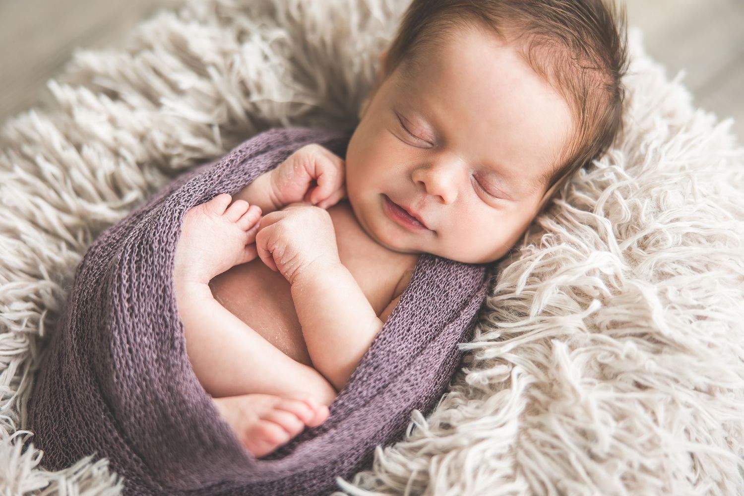 Sleeping newborn baby wrapped in purple knit blanket on fluffy white rug.