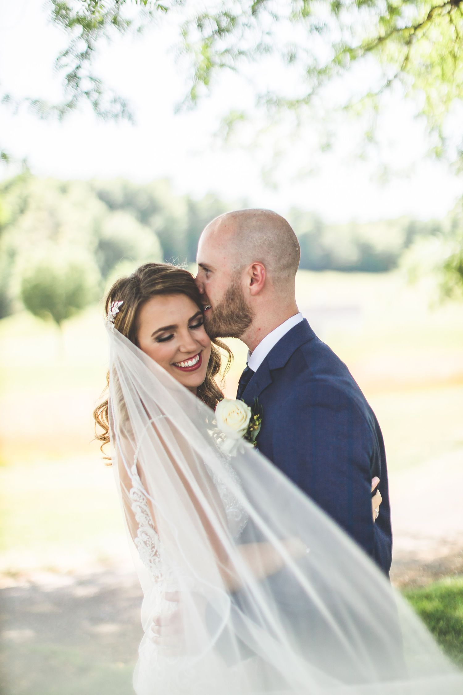 An intimate wedding portrait captured outdoors with a flowing veil in natural sunlight.