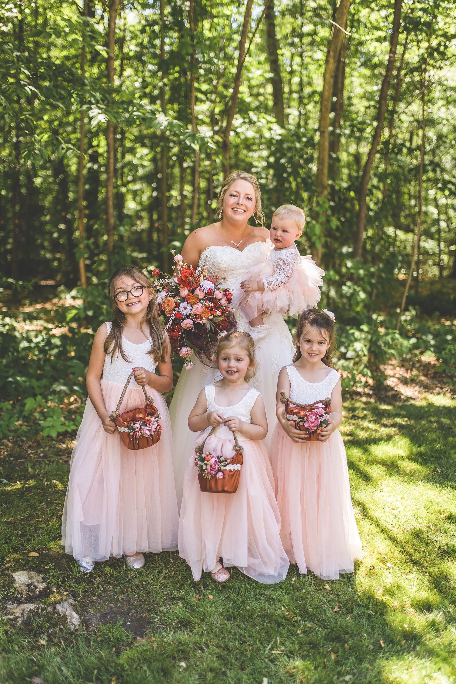 Young attendants in pink dresses carry flower baskets through a sunlit wooded area.