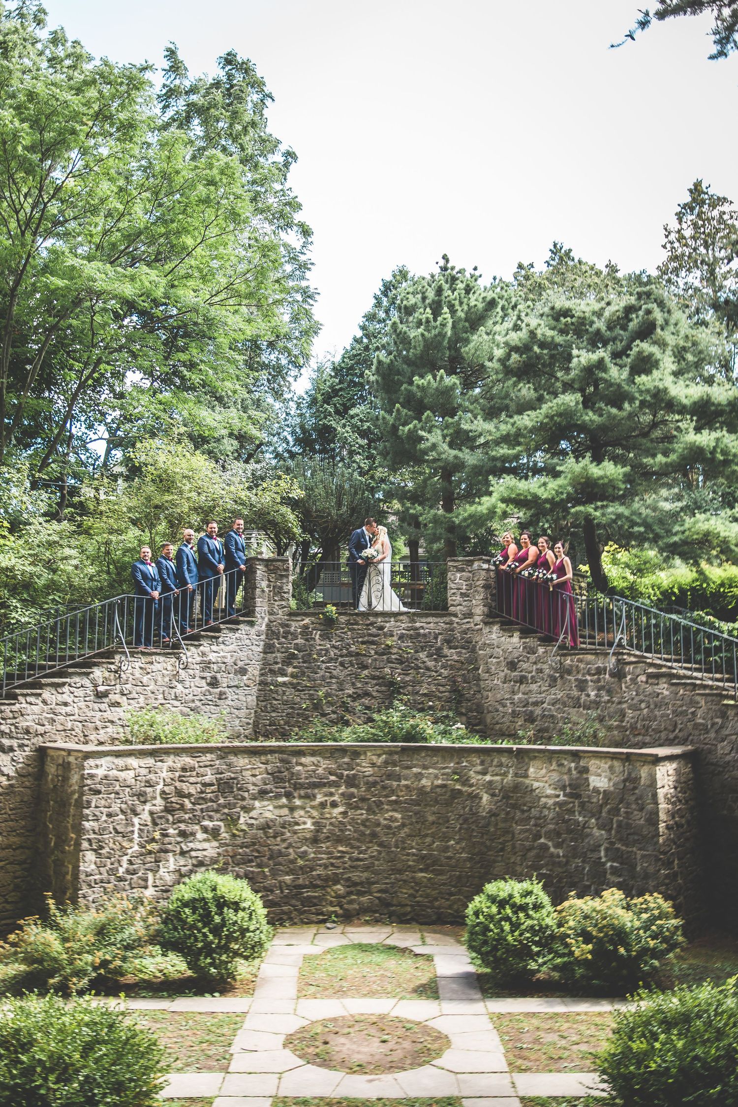 Wedding party posing on a rustic stone wall surrounded by green trees.